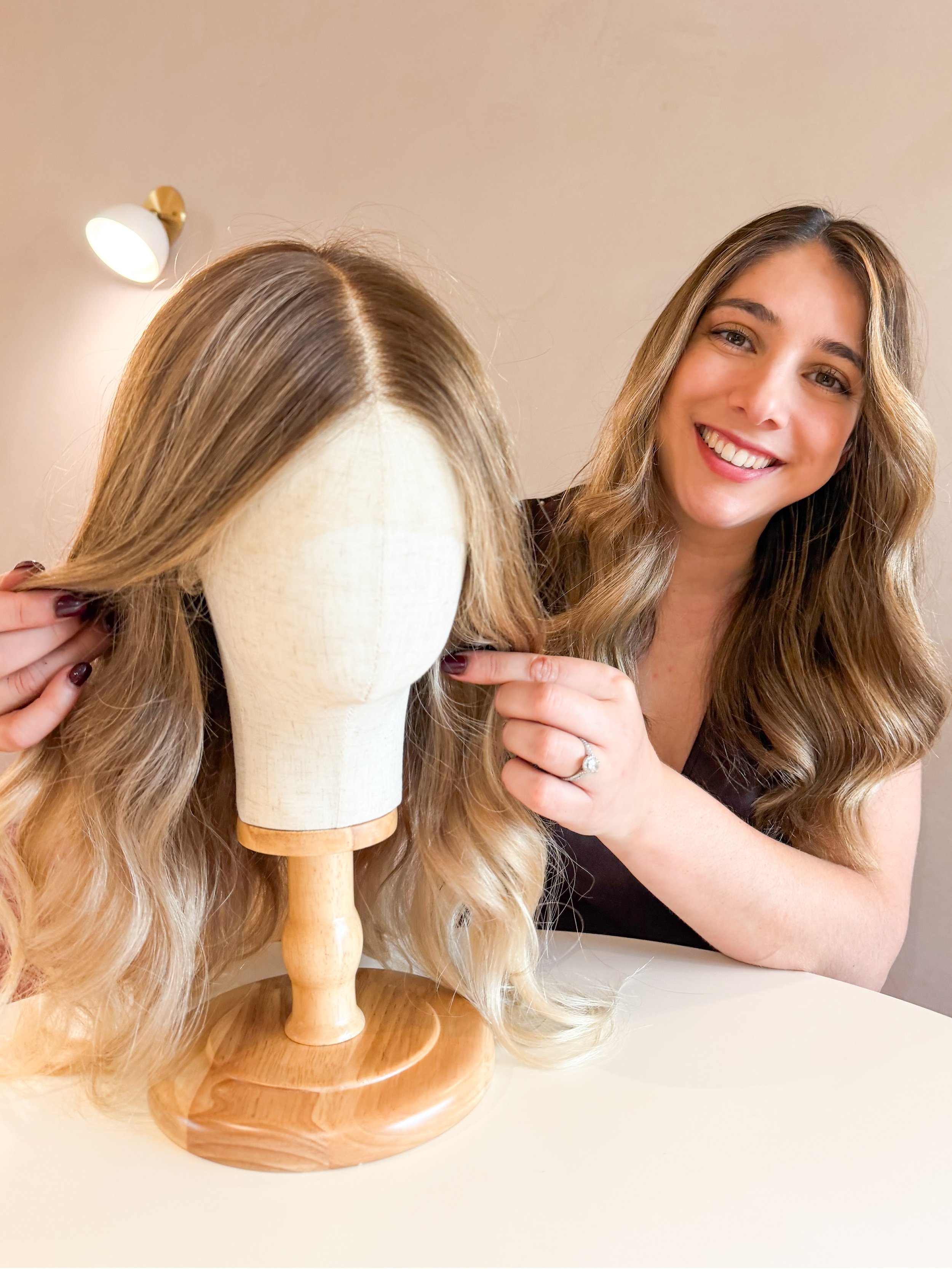 A woman with long, wavy hair smiling at the camera, pointing at a blonde wig on a mannequin with a wooden stand, in a room with a wall LED light in the background.
