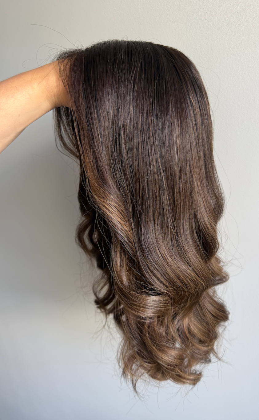 A hand holding a wig with wavy brown hair against a plain white wall