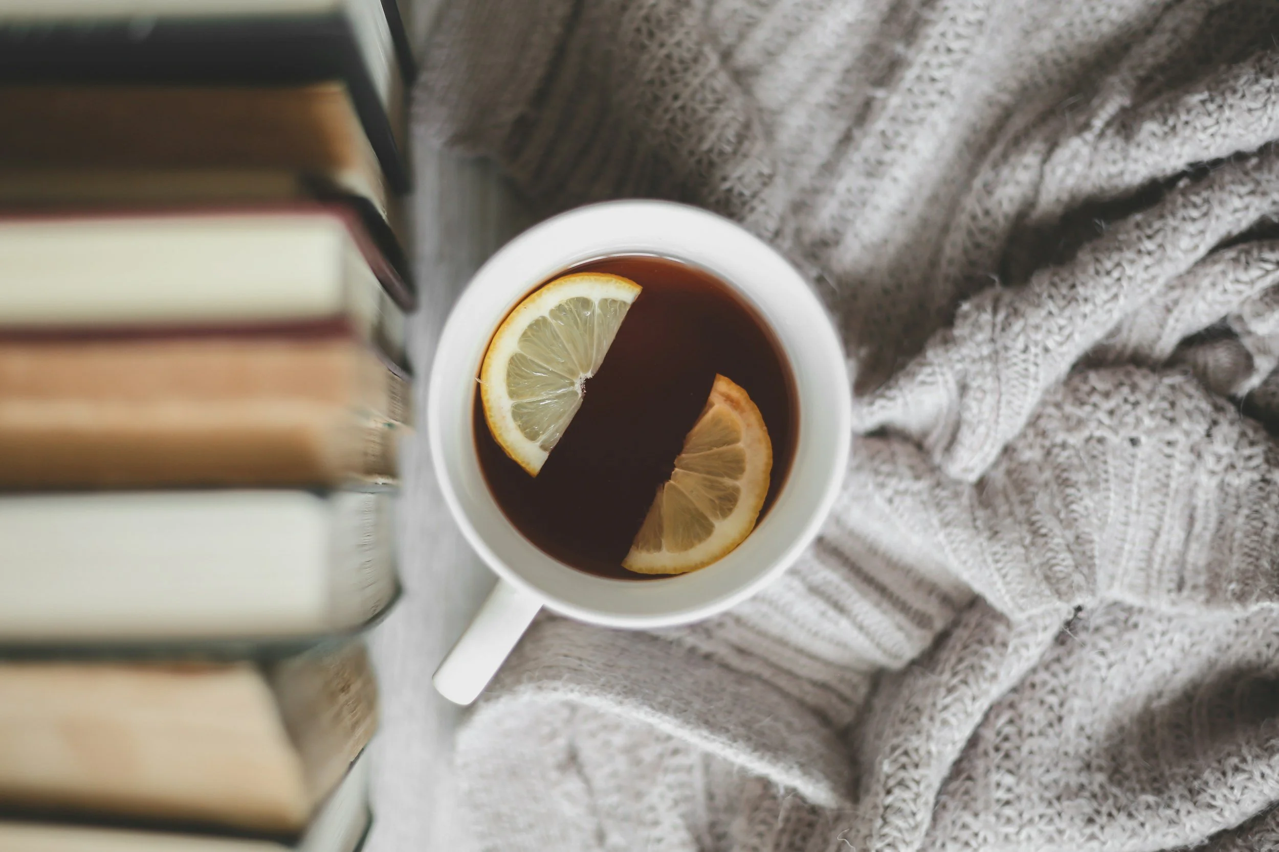 A white mug of black tea with lemon slices, resting on a cozy gray knit blanket, next to a stack of books.