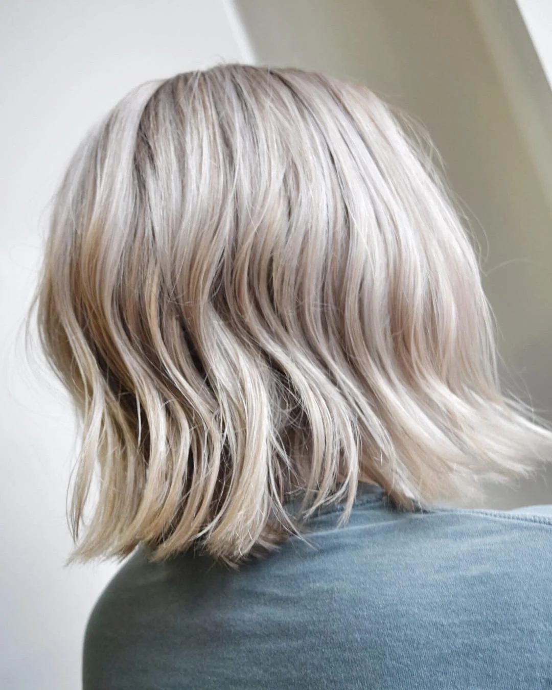 Side view of a person with shoulder-length, wavy platinum blonde hair, sitting against a neutral background.