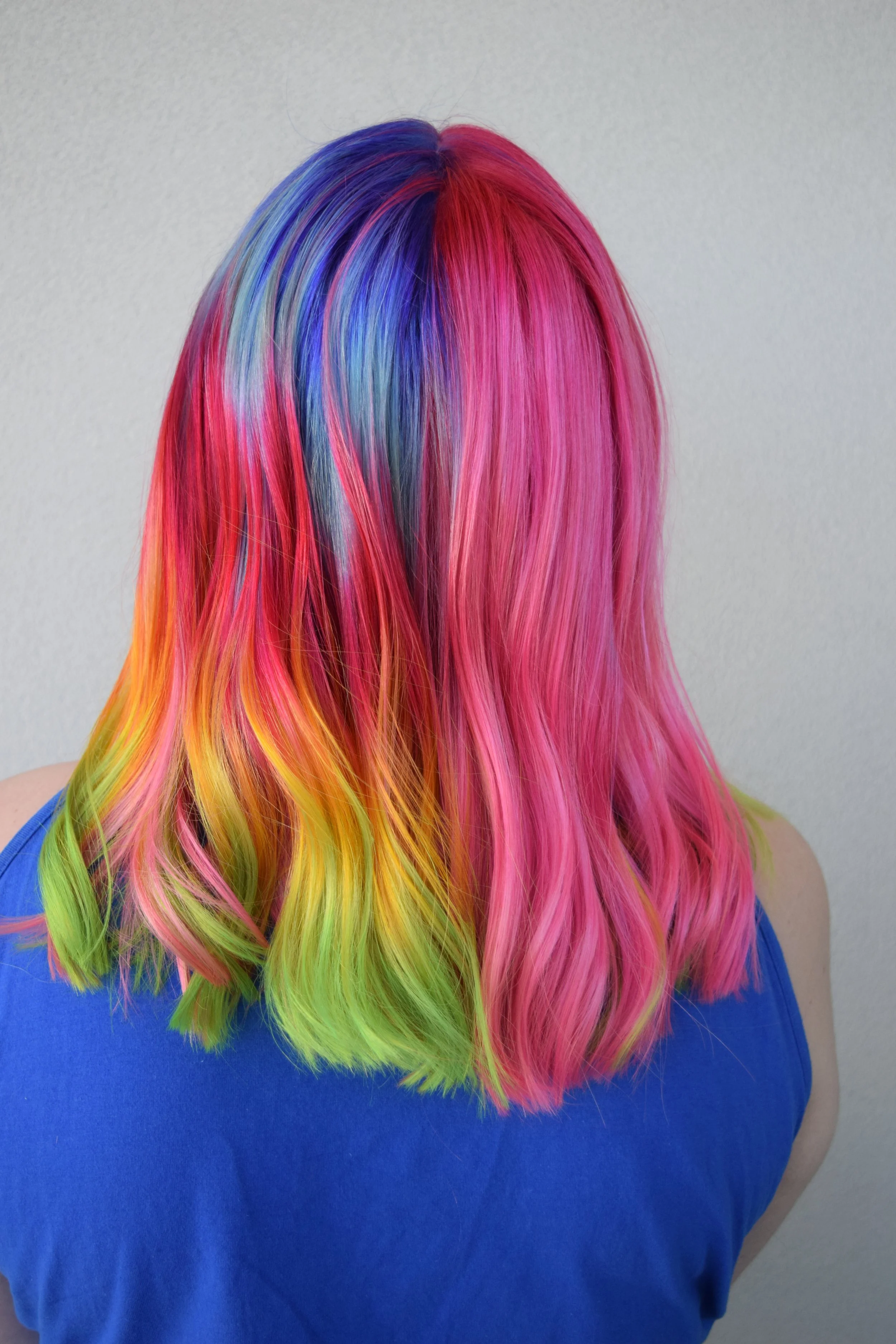 Back view of a person with vibrant, multi-colored rainbow hair, wearing a blue sleeveless top, standing against a plain light-colored wall.