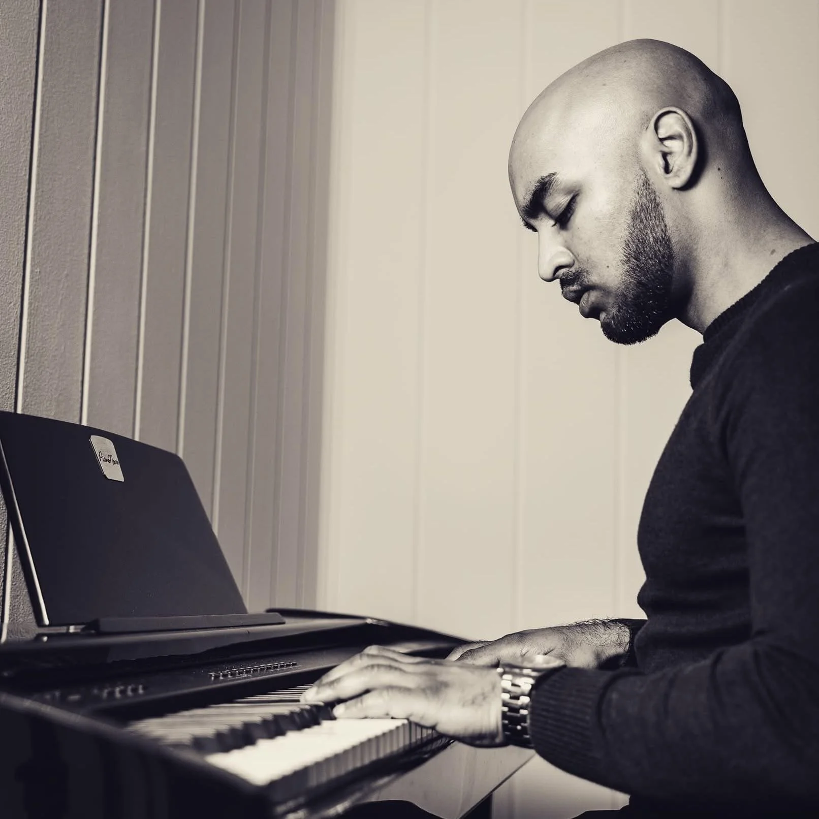 OG Vibez speelt piano in een kamer met beige houten wanden, afgebeeld in zwart-wit.