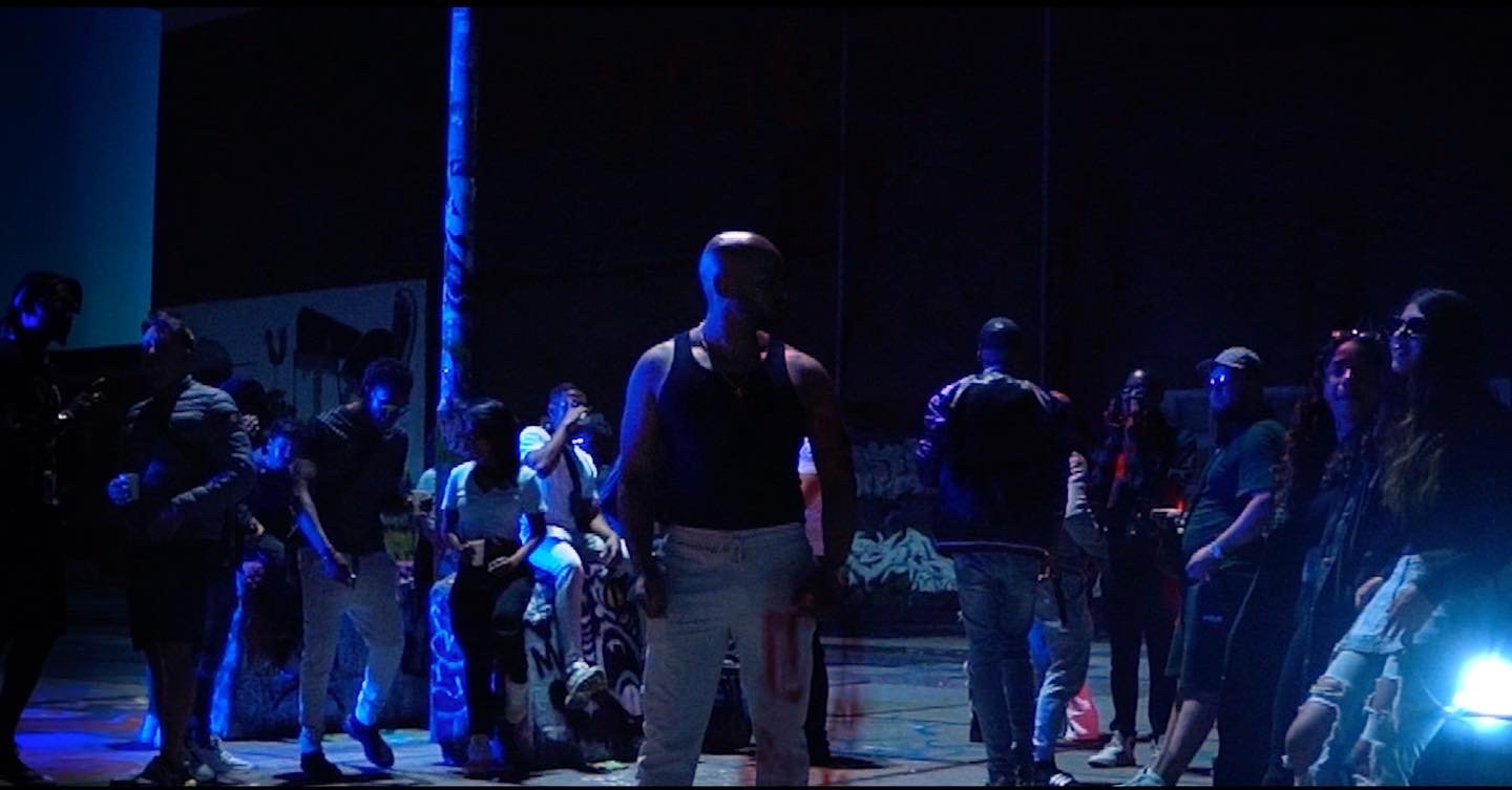 OG Vibez clipshoot, mensen staan in een donkere ruimte met blauwe verlichting, sommige mensen dragen masks en casual kleding.