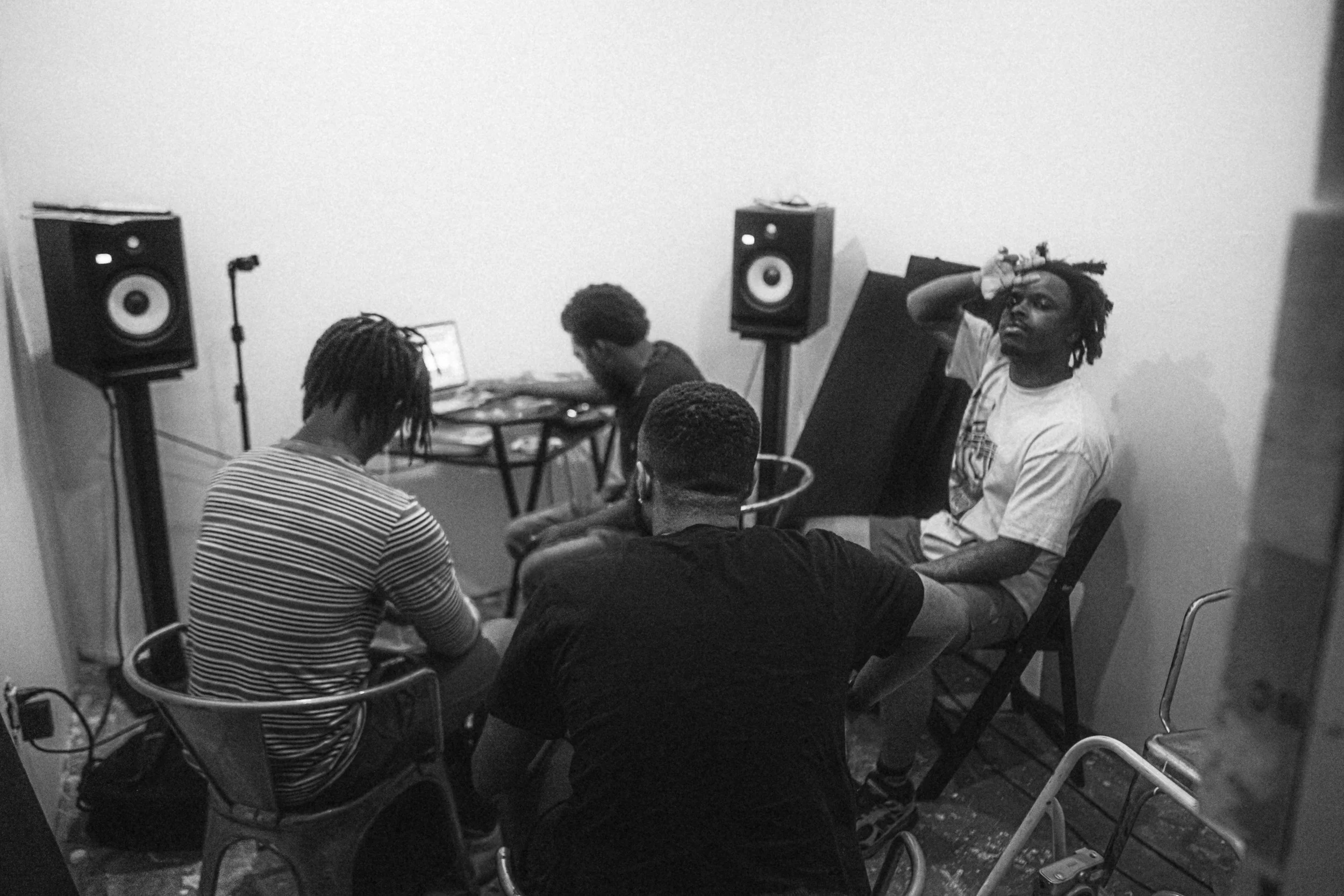 Vier mensen in een opnamestudio, twee zijn aan het werken met geluidsapparatuur en twee zitten in stoelen, één met hand op hoofd. Workshop met OG Vibez