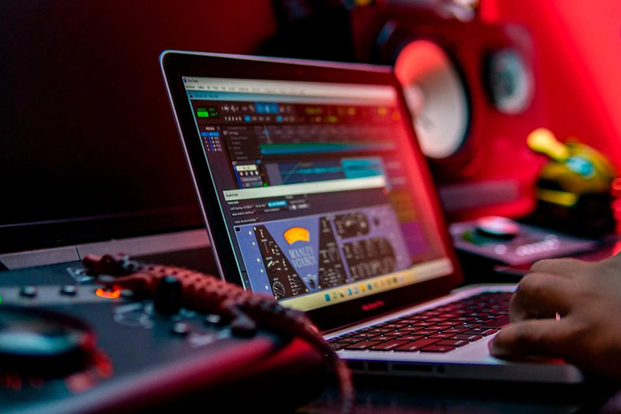 Close-up van een laptop met muziekproductiesoftware op het scherm, omringd door audio apparatuur en een hand die op het toetsenbord drukt, met rode achtergrondverlichting. OG Vibez die een beat maakt