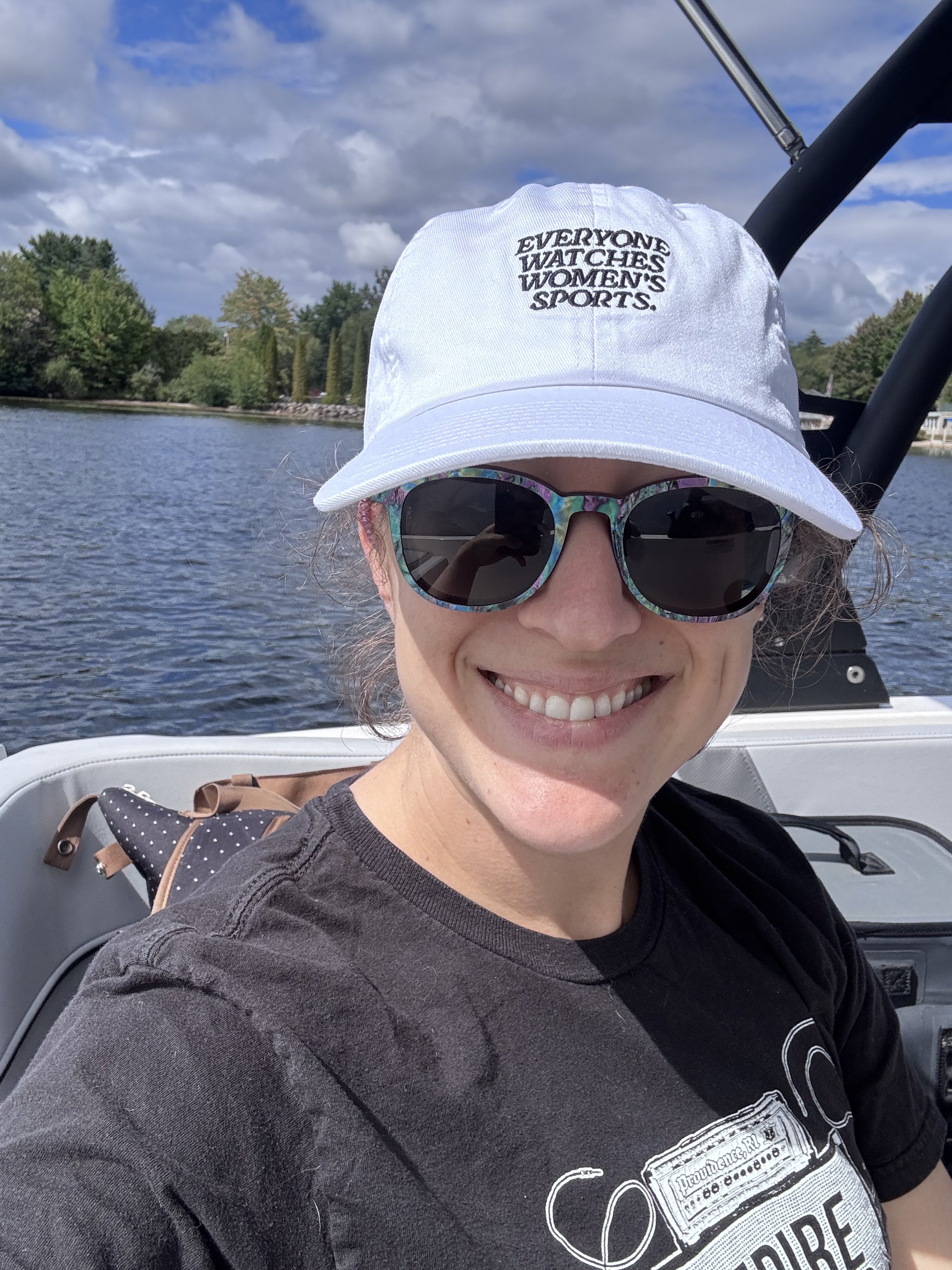 Selfie of lauren on a boat in a lake, wearing a black T-shirt, multi-colored sunglasses, and white hat that reads "Everyone Watches Women's Sports." in black letters.