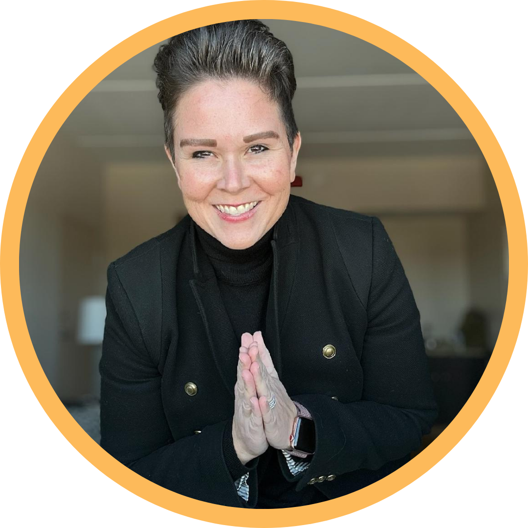 Woman smiling with hands in a prayer position, wearing a black jacket and a black turtleneck, indoors with a blurred background.