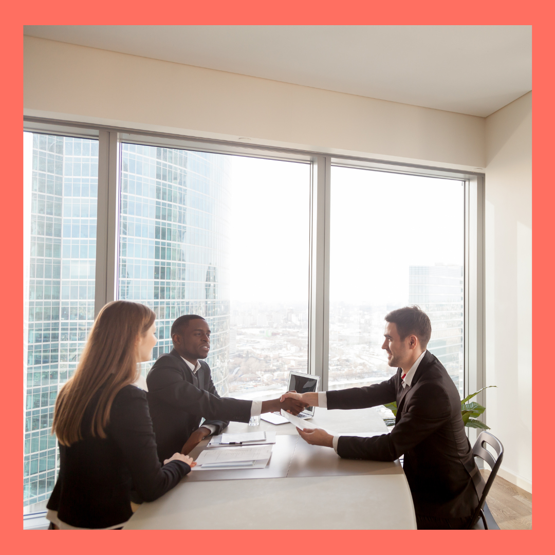 Three business professionals in a modern office, two men and one woman, are engaged in a handshake over a conference table during a business meeting, with large windows revealing a cityscape of tall glass buildings in the background.