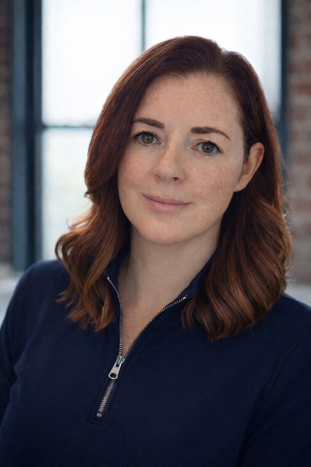 Close-up of a smiling woman with shoulder-length reddish-brown hair, wearing a navy blue zip-up jacket, in an indoor setting with large windows and brick walls.