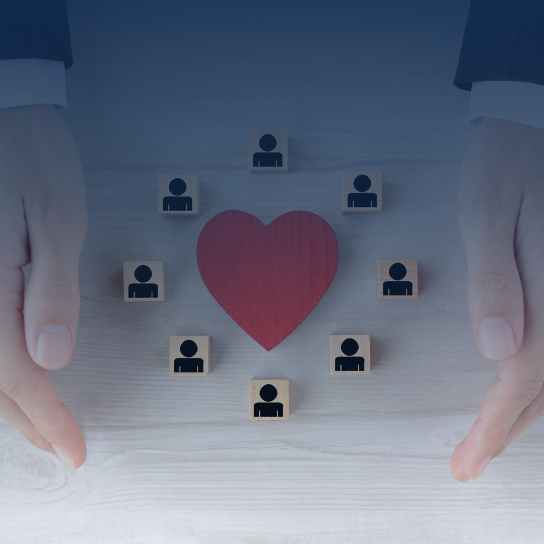 Hands surrounding a red heart and small icons of people on a light wooden surface.