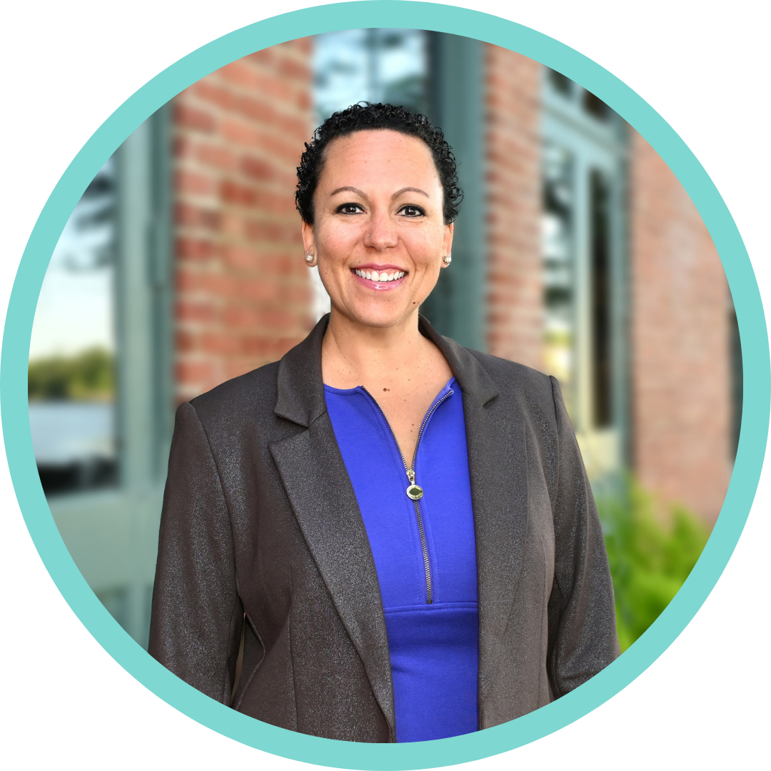 A woman with short curly black hair smiling outdoors, wearing a dark blazer over a blue zip-up top, standing in front of a building with large windows and brick walls.