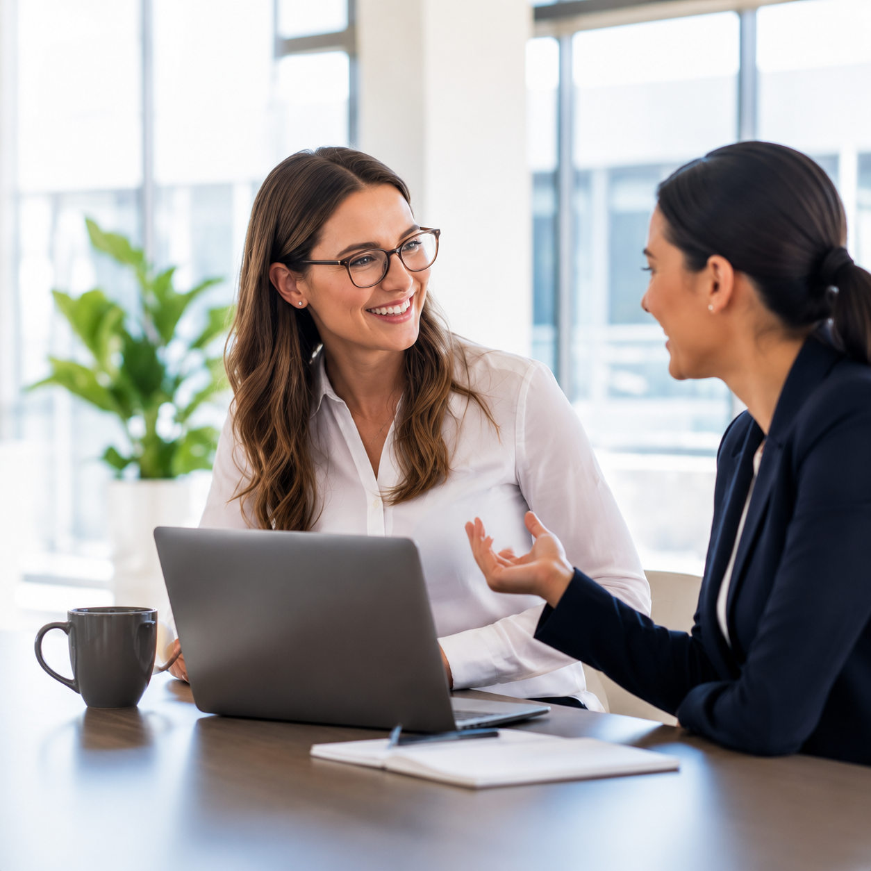 Two women having a discussion in an office, smiling at each other, with a laptop, coffee mug, and notebook on the table.