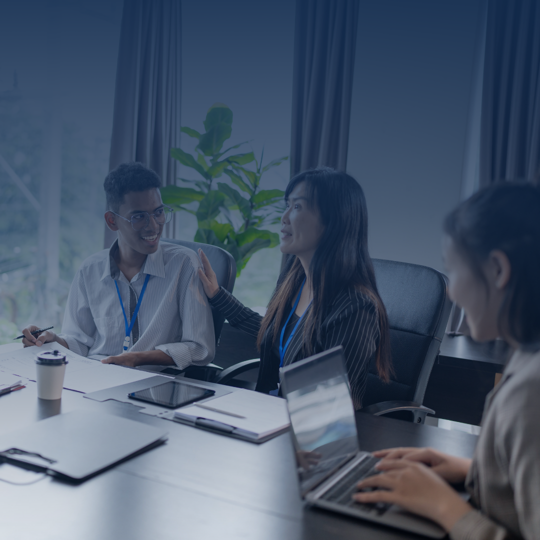 Four diverse people sitting at a conference table in a meeting room, engaged in a discussion. One woman is speaking and gesturing, while another woman is typing on a laptop. The room has large windows, curtains, and indoor plants.