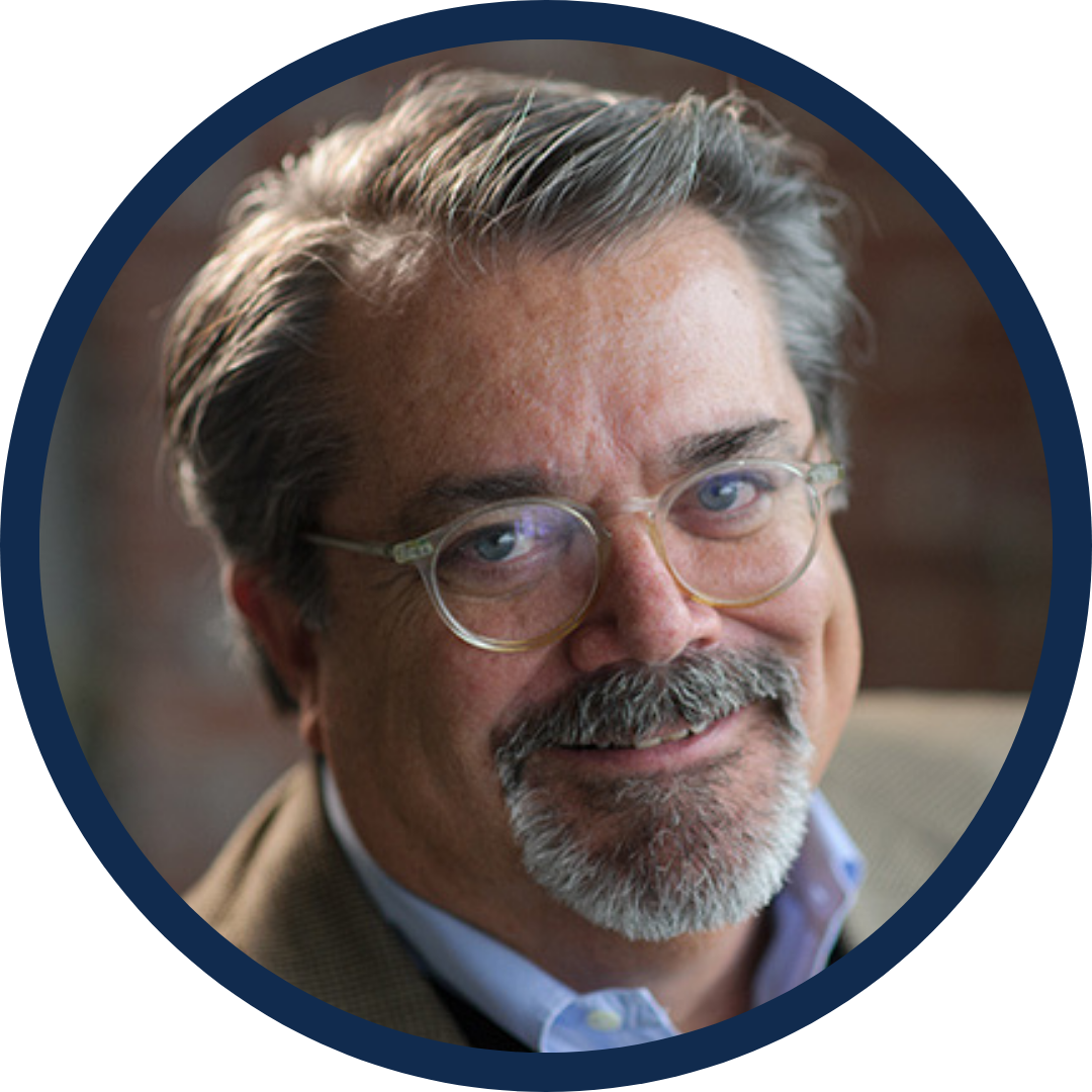 Close-up portrait of a middle-aged man with gray hair, beard, and glasses, smiling gently indoors.