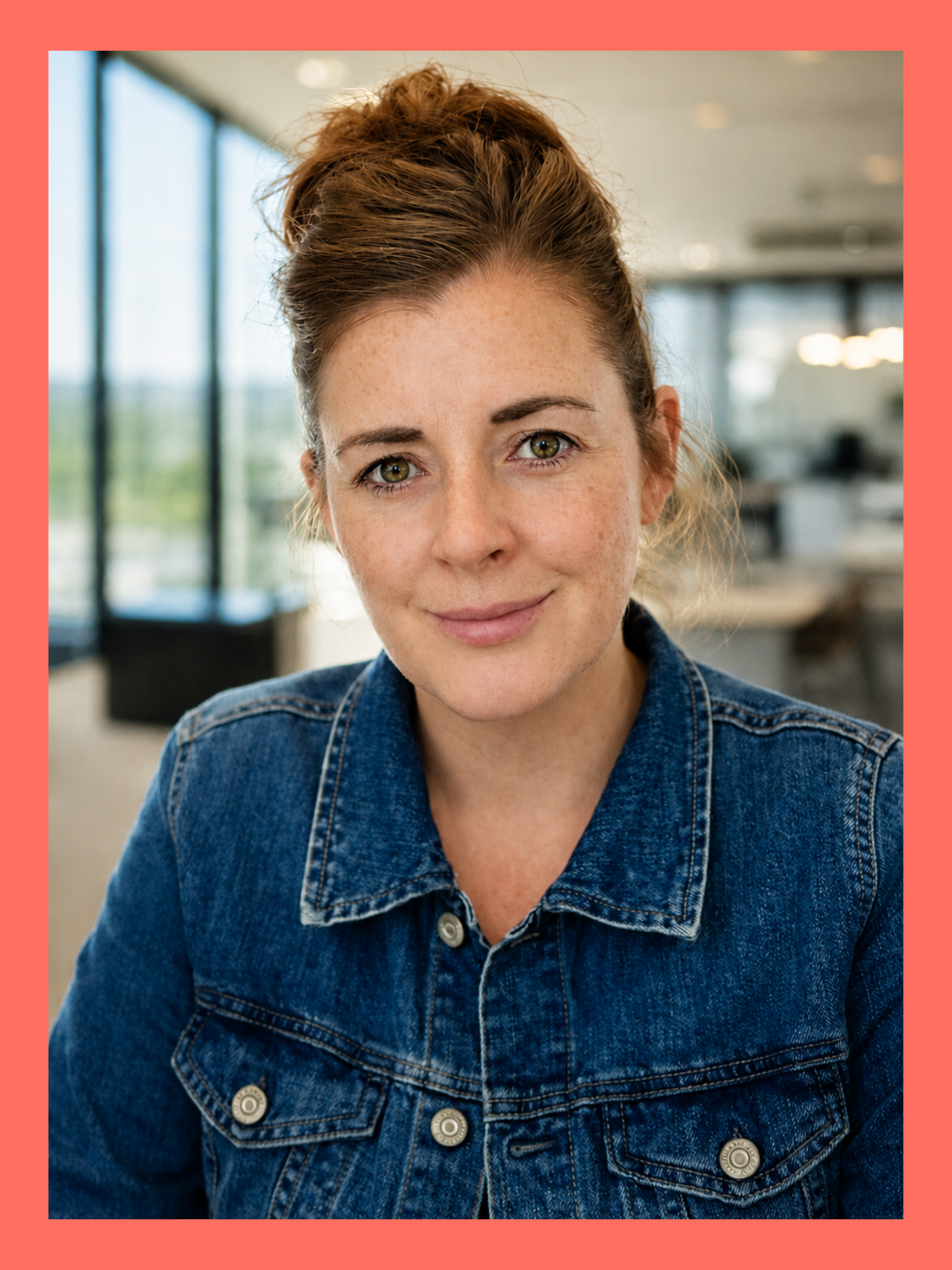A young woman with a bun hairstyle and light freckles, wearing a denim jacket, smiling in a bright indoor setting with large windows.