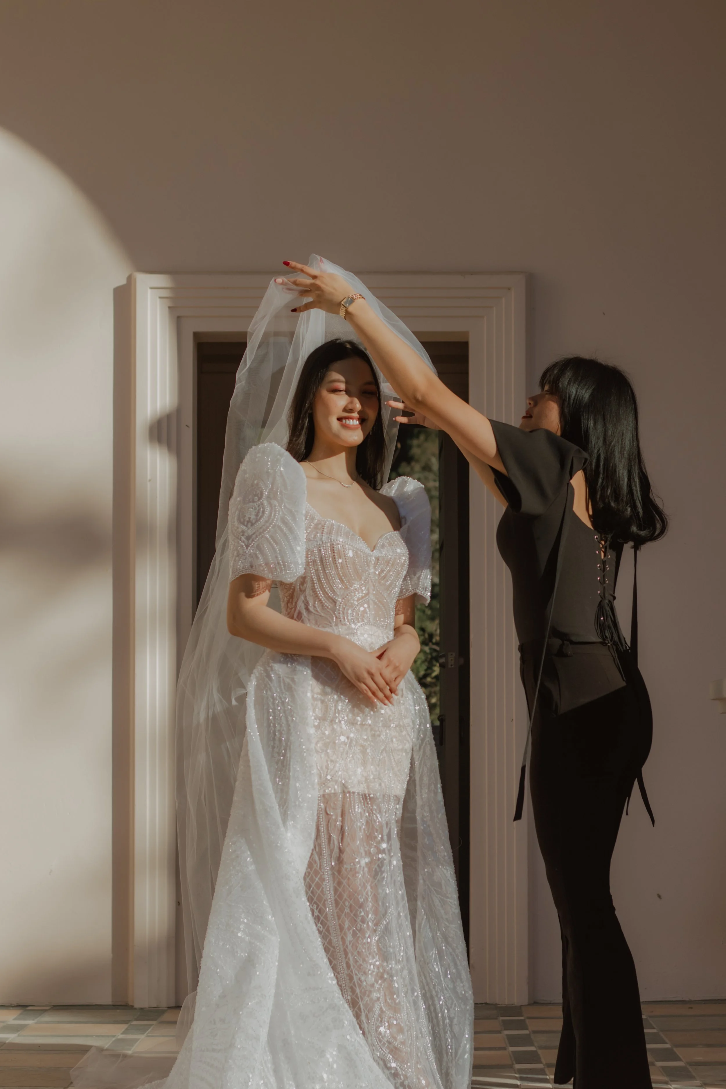 A woman in a wedding dress smiling as another woman adjusts her veil inside a room.