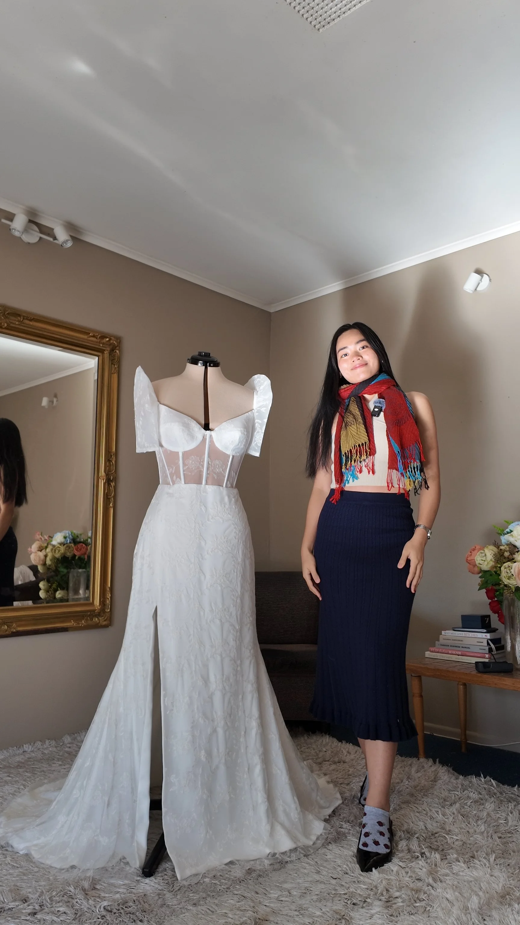 Woman standing next to a wedding dress on a mannequin in a room with beige walls, a mirror, a side table with flowers and books.