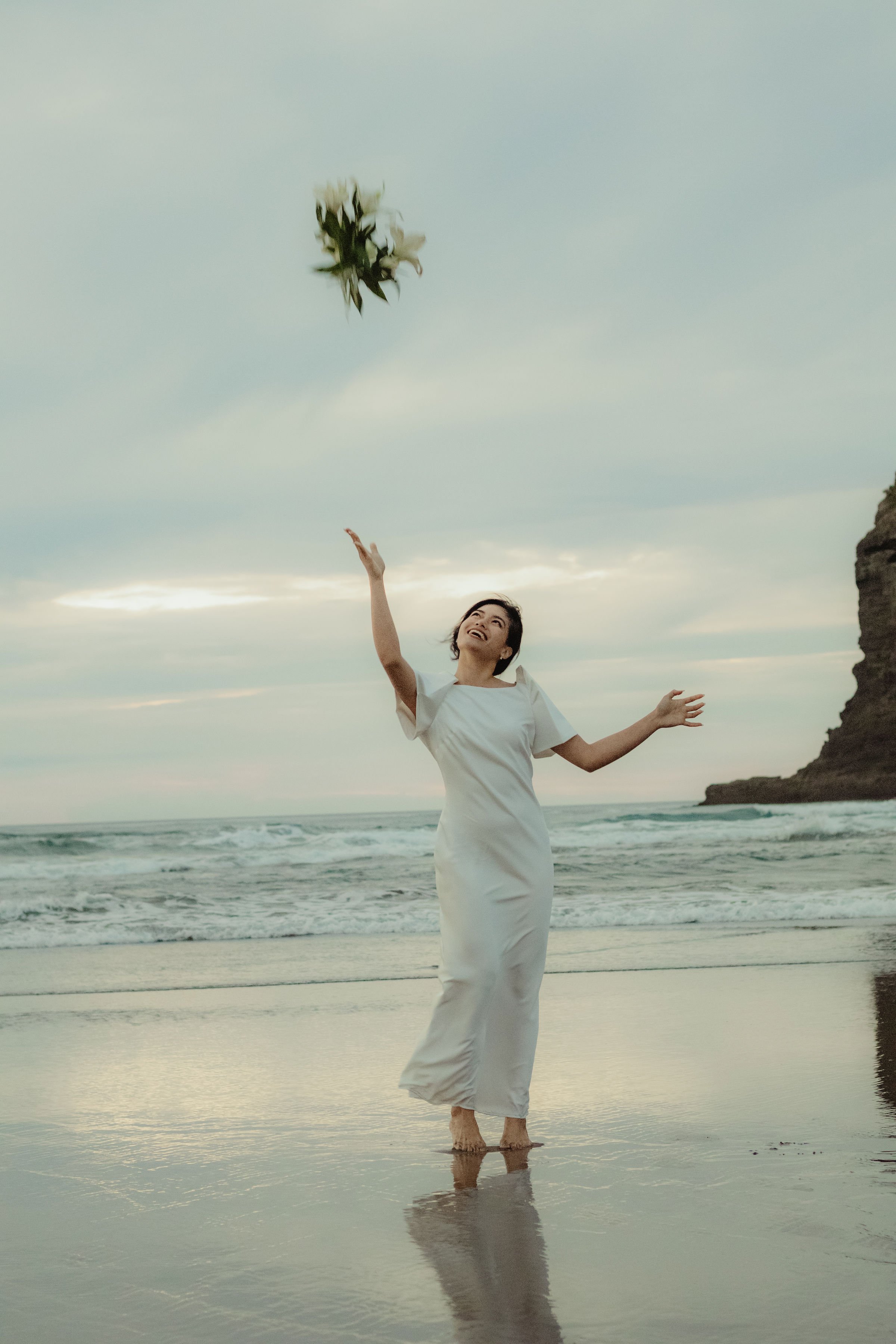 A woman in a white dress standing barefoot in shallow water at the beach, throwing a bouquet of flowers into the air and smiling, with ocean waves and a cloudy sky in the background.