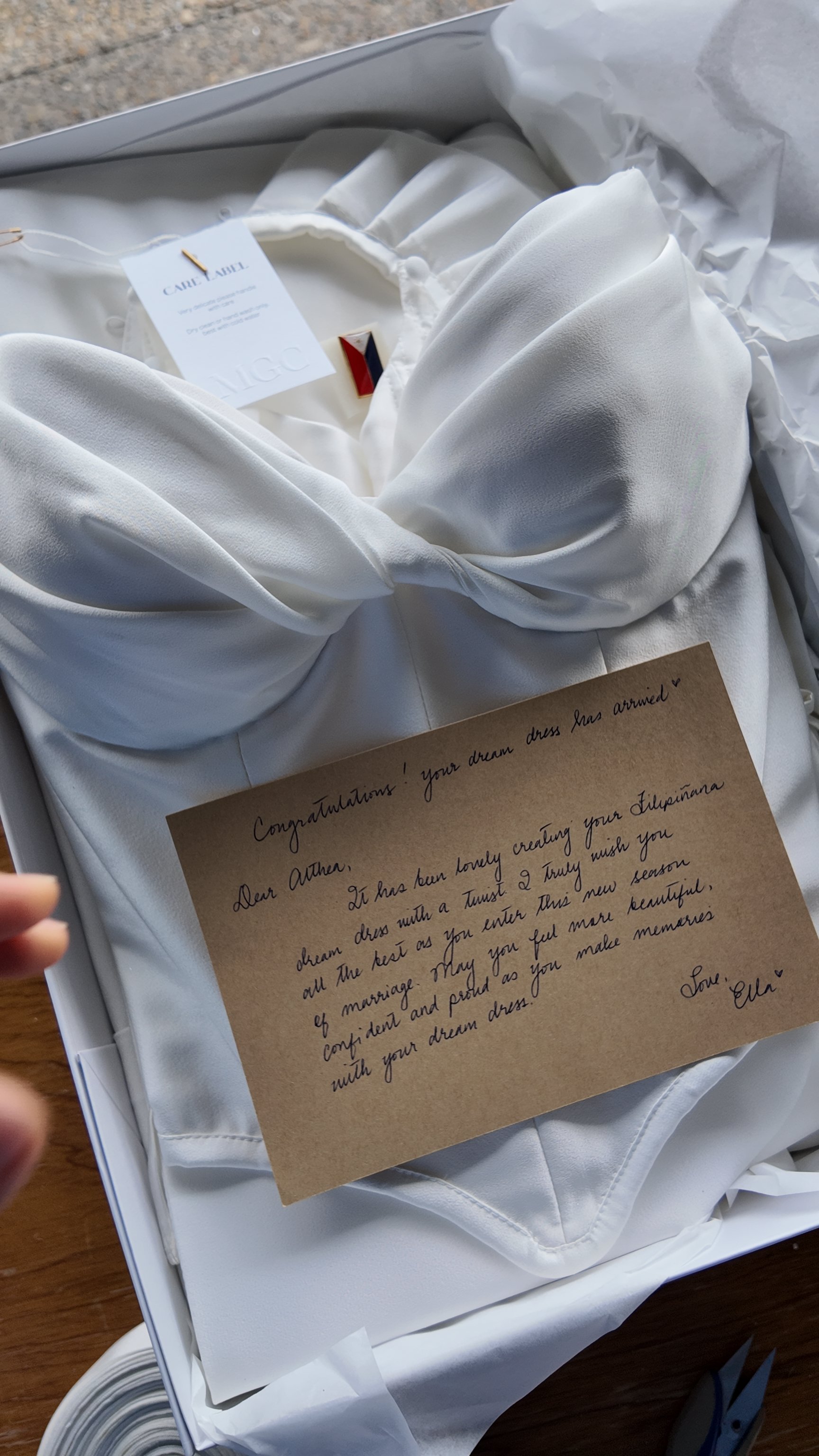 A white wedding dress inside a box with a handwritten note, a glow necklace, and a small decorative item. The note congratulates the recipient on their wedding dress.