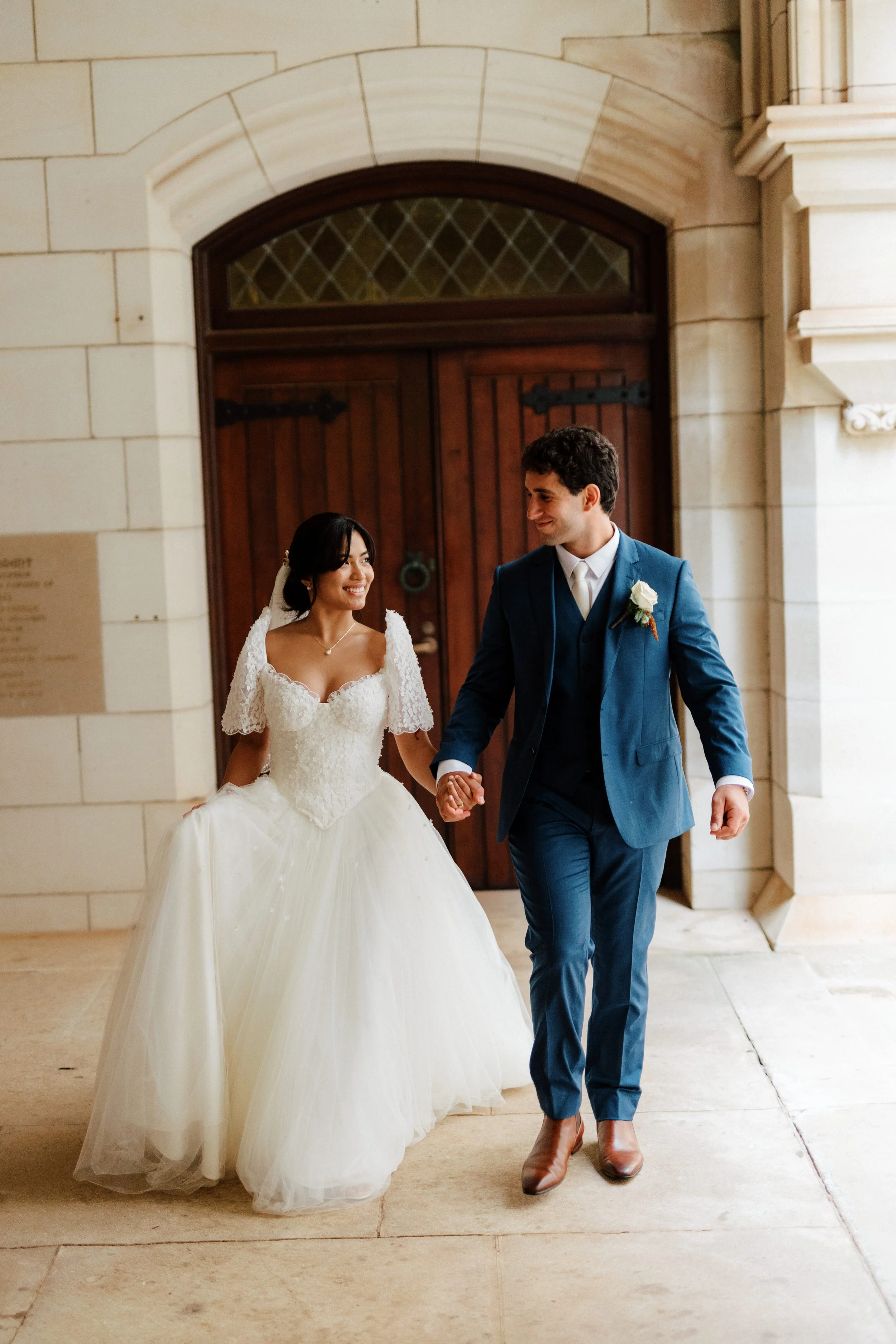 A bride and groom holding hands and smiling walking together in a hallway with a large wooden door behind them.