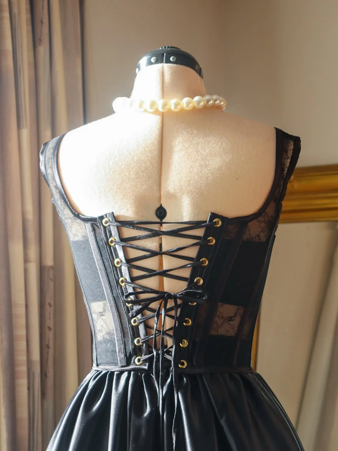 Black corset with lace details and gold eyelets on a dress form, draped in a pearl necklace and black satin skirt with pleats, set against a background with a beige wall and window with curtains.