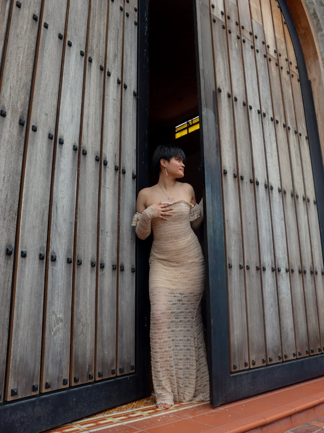A woman in an off-the-shoulder, beige, lace gown stands in the doorway of a wood-paneled building, looking to her left.