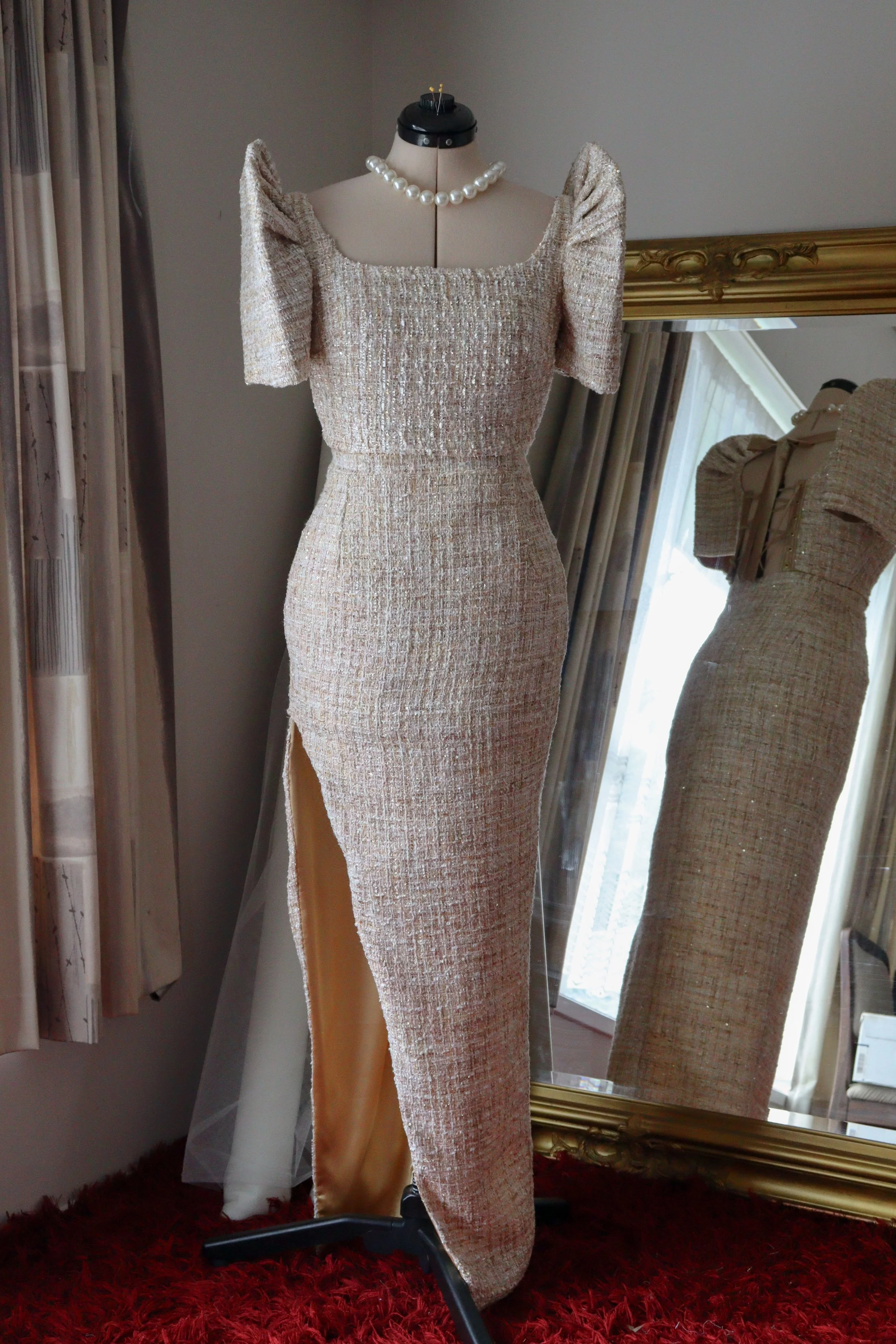 A mannequin dressed in a beige, textured, elegant evening gown with puffed sleeves and a thigh-high slit, accessorized with a pearl necklace, standing in front of a large gold-framed mirror.