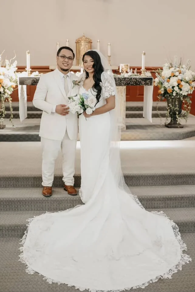 A bride and groom standing together inside a church, smiling, with a wedding altar decorated with flowers and candles behind them.