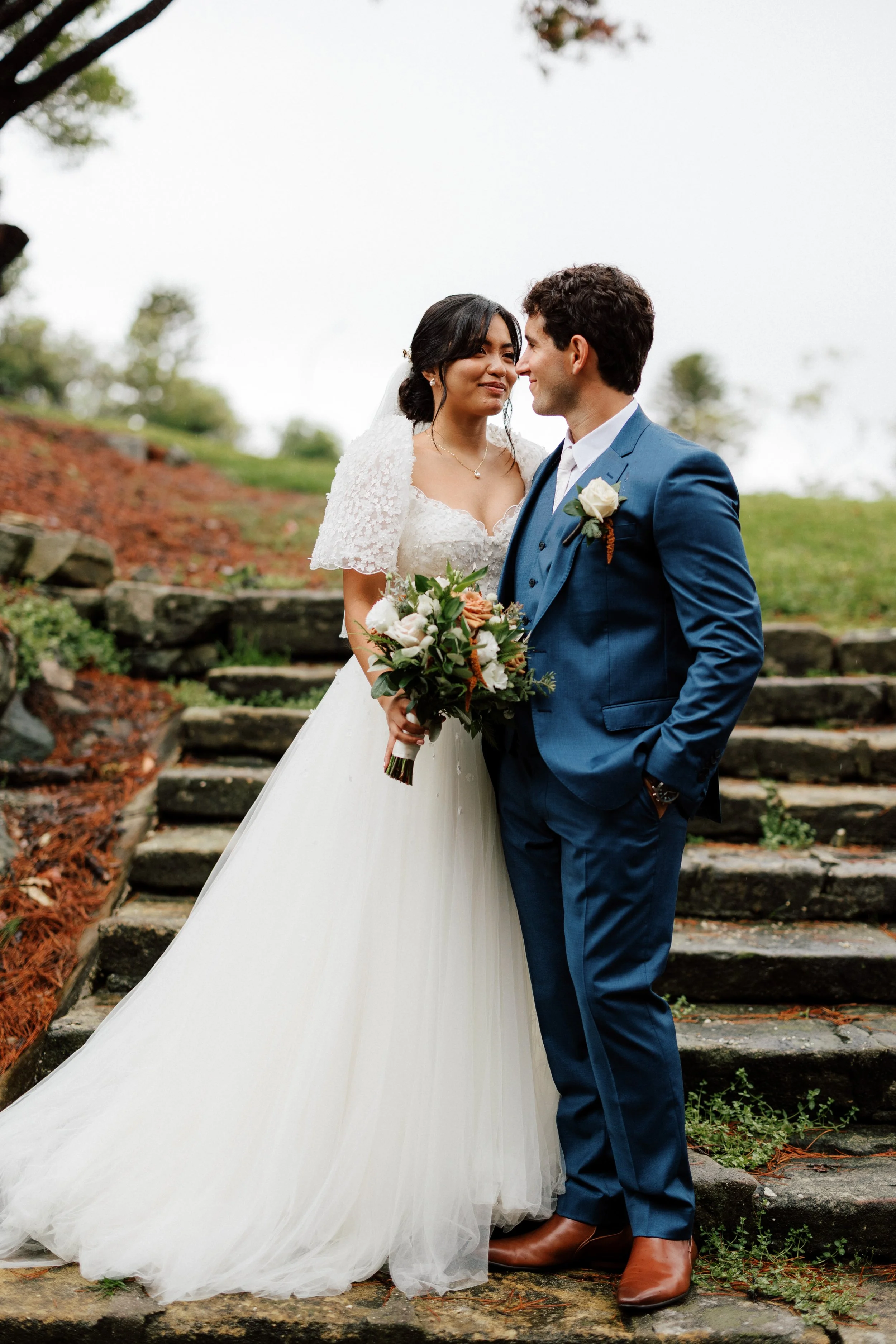 A bride and groom stand close together outdoors on stone steps. The bride wears a white lace wedding gown and holds a bouquet of flowers. The groom wears a blue suit with a white shirt and a white boutonniere. They look into each other's eyes with a backdrop of greenery and a cloudy sky.