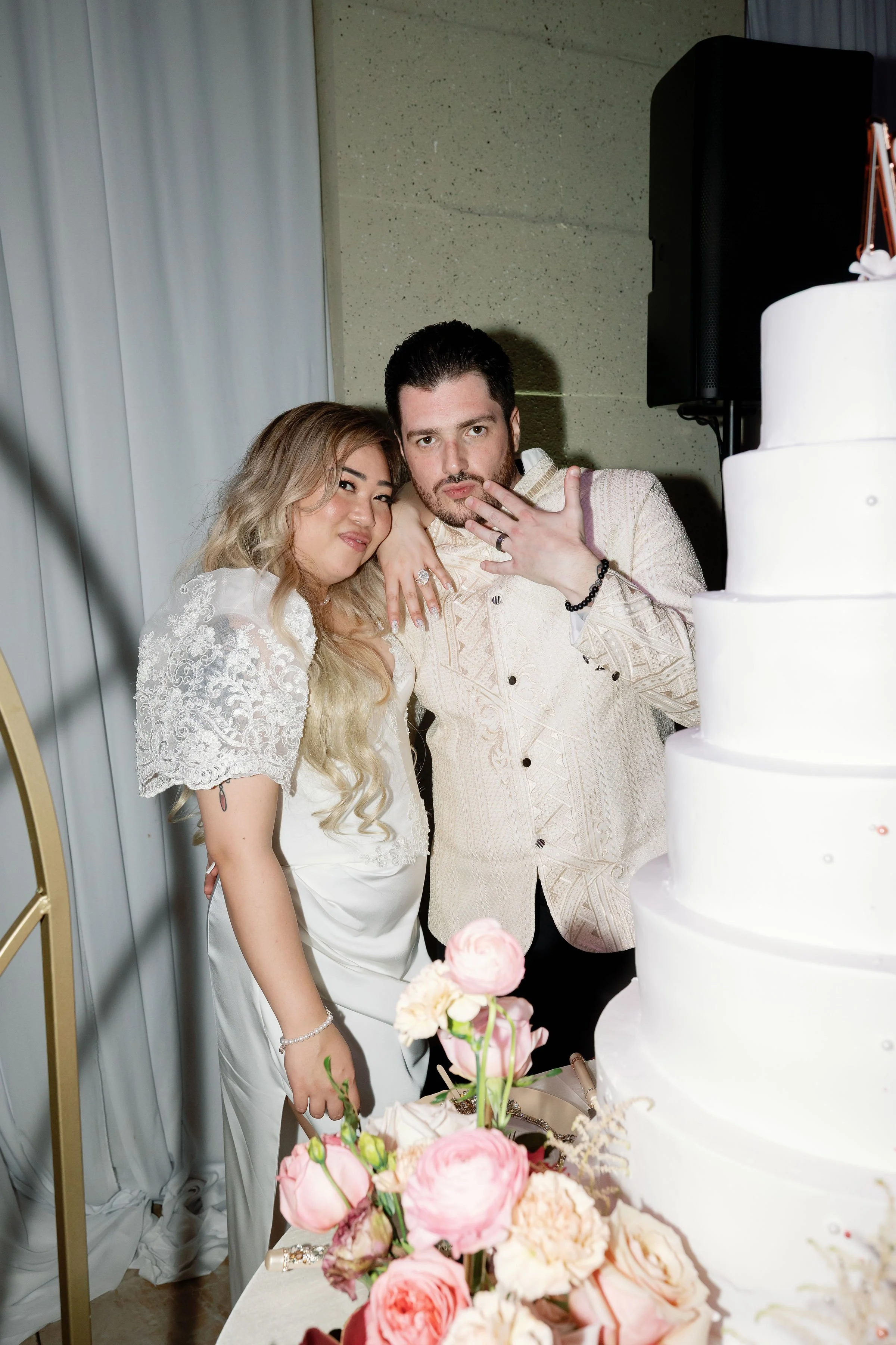A couple dressed in wedding attire posing together at their wedding reception, with a large white wedding cake and pink flowers nearby.