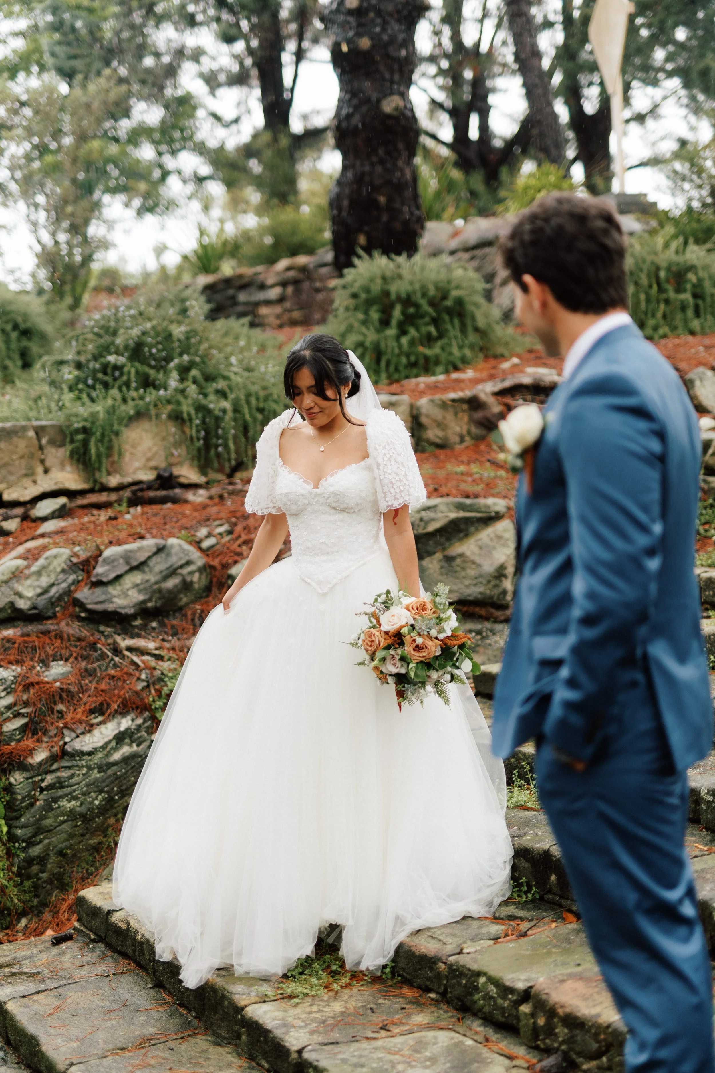 Filipina Bride in white wedding dress holding bouquet looking down as groom in blue suit watches at outdoor wedding.