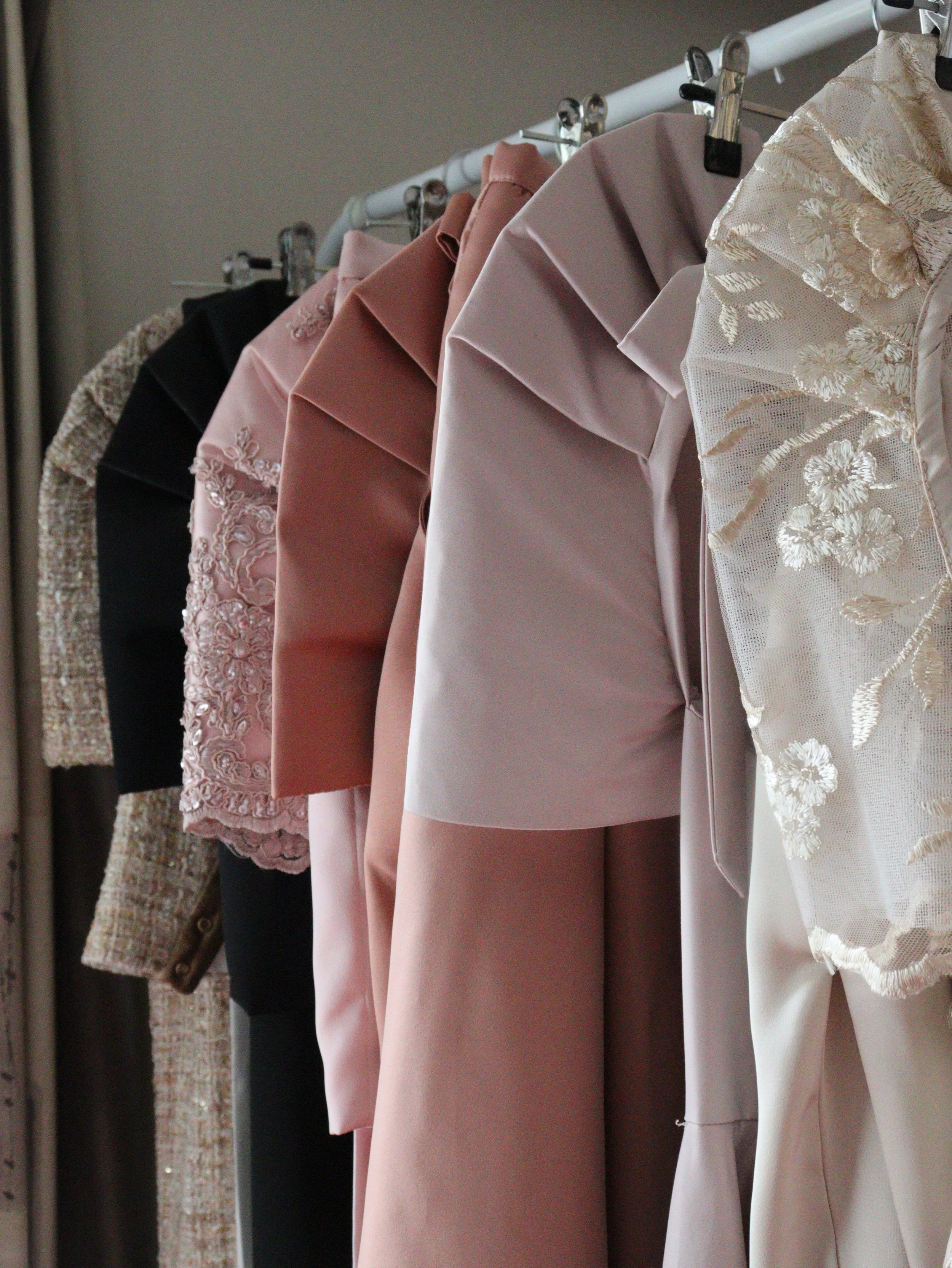 Various formal dresses, including blush pink, black, and lace with floral embroidery, hanging on a clothing rack.
