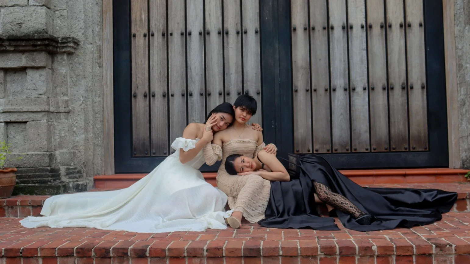 Three women dressed in elegant gowns sitting on brick steps in front of a large wooden door, posing closely together.