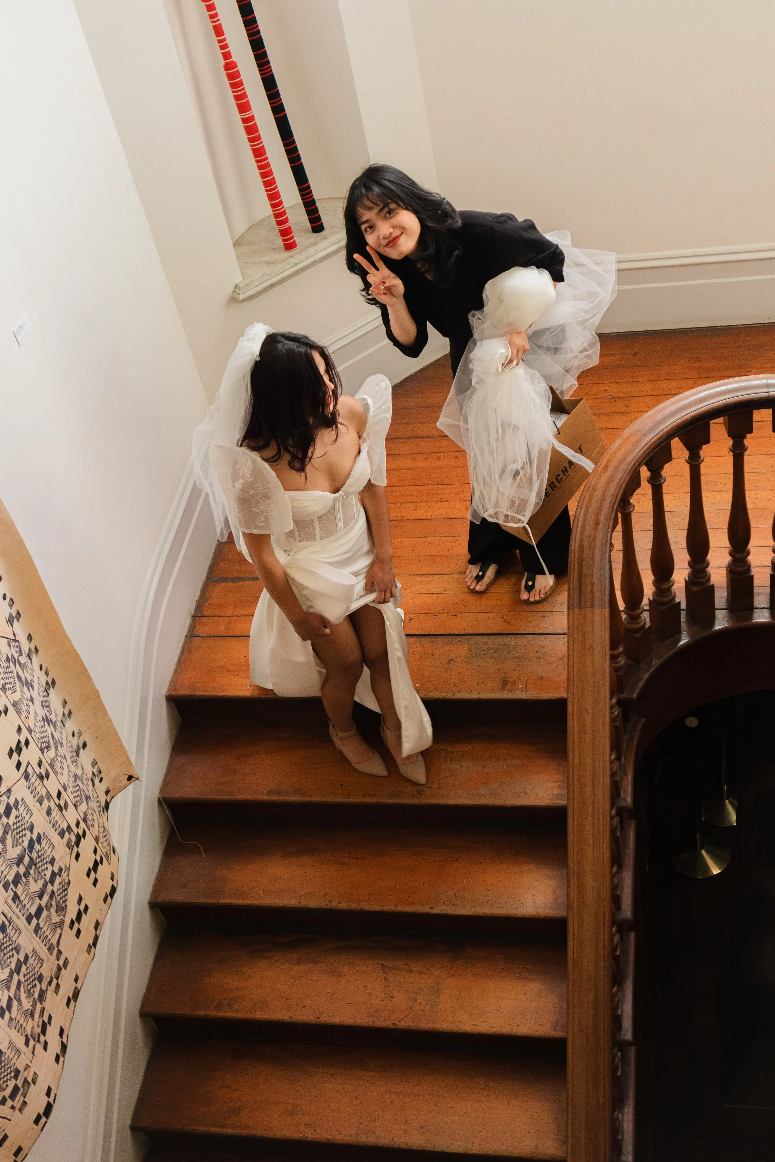 Two women on a wooden staircase, one in a wedding dress and veil, and the other in black clothing making a peace sign, holding balloons and a gift bag.