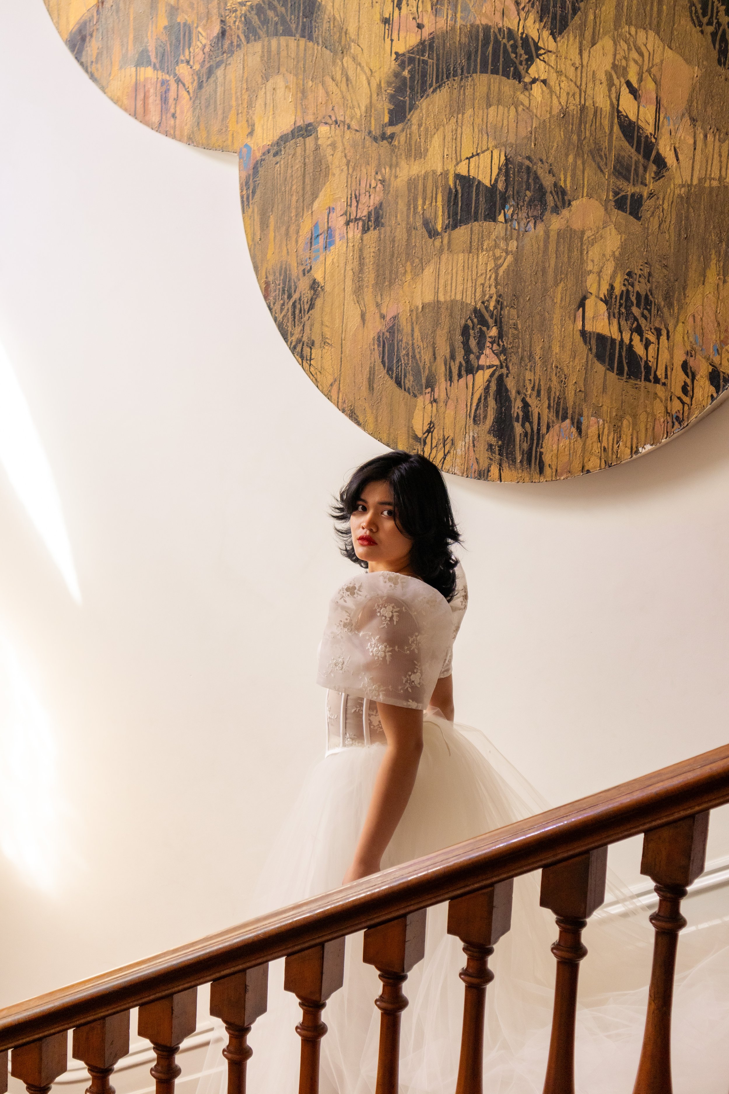A woman in a white dress standing on a staircase, looking over her shoulder, with a large abstract gold and black painting on the wall behind her.