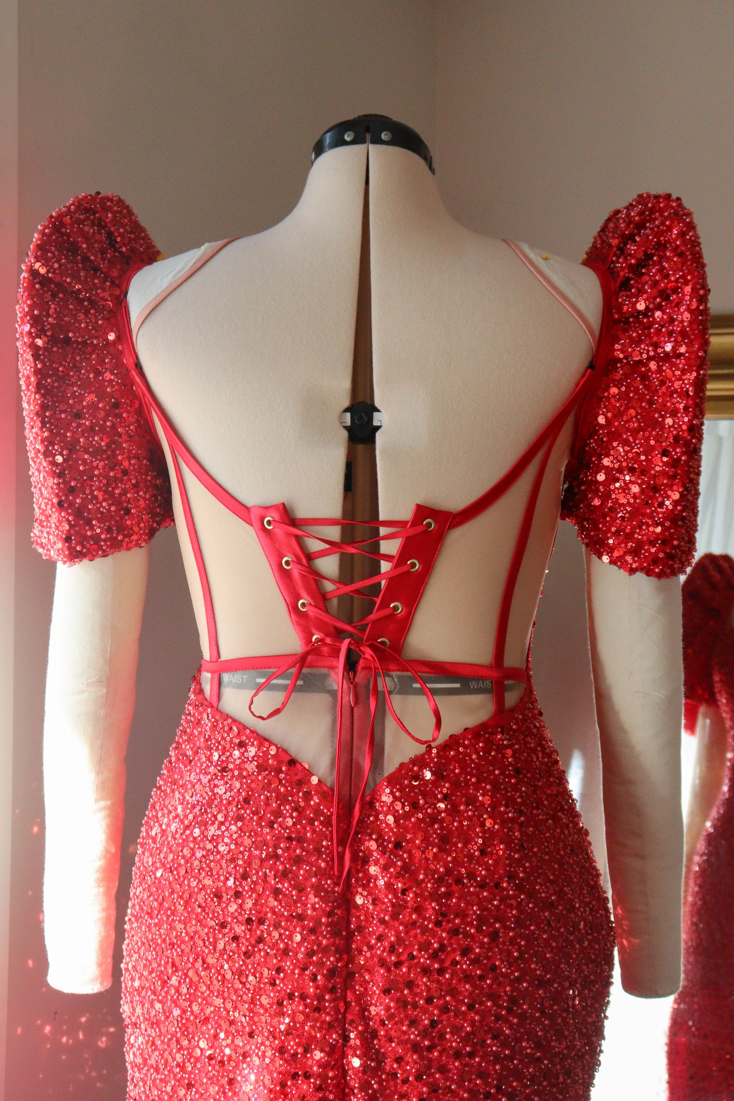 Red sequined gown with lace-up back and sheer panels displayed on a mannequin.