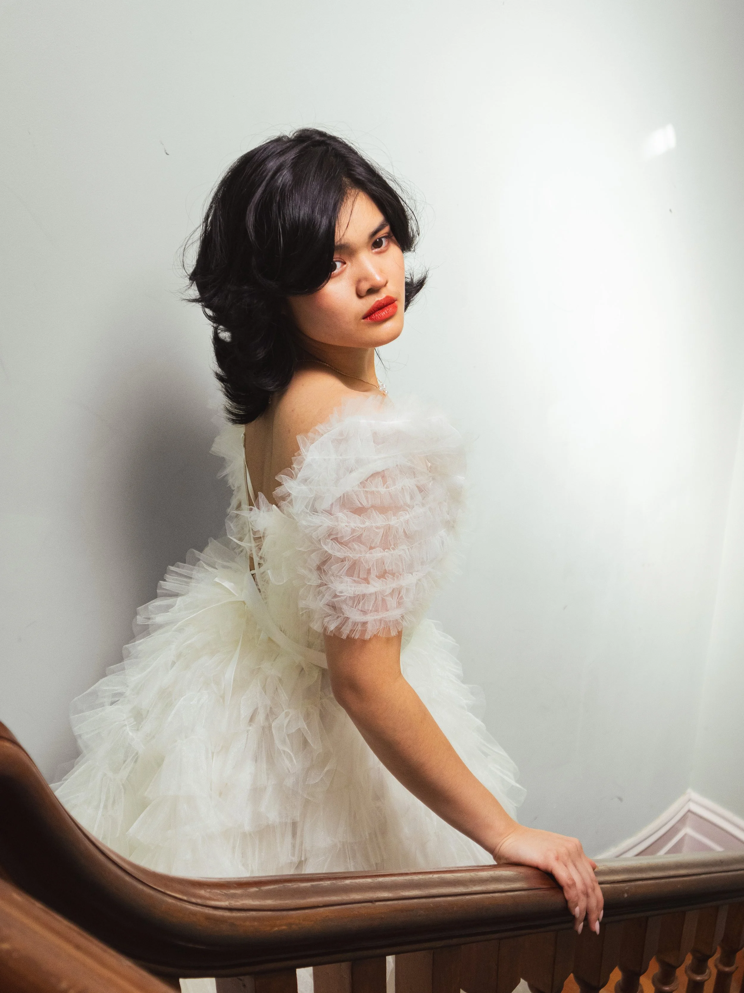 A woman with dark, wavy hair and red lipstick wearing an elegant white, ruffled dress standing on a staircase with wooden railing, looking at the camera.