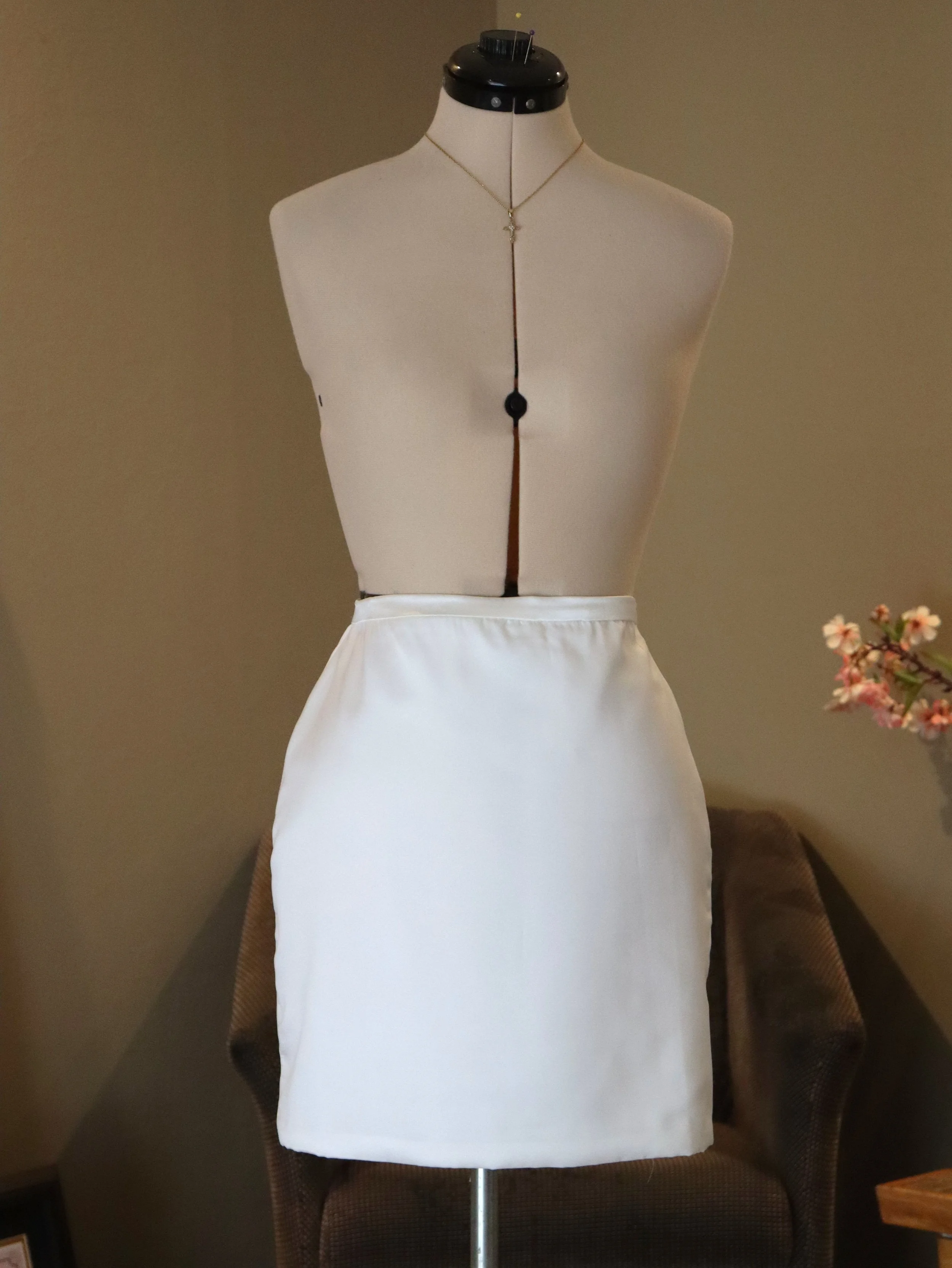 A dress form wearing a necklace and a white apron, standing in a room with a beige wall, a dark brown chair, and a small pink flower arrangement on a table.