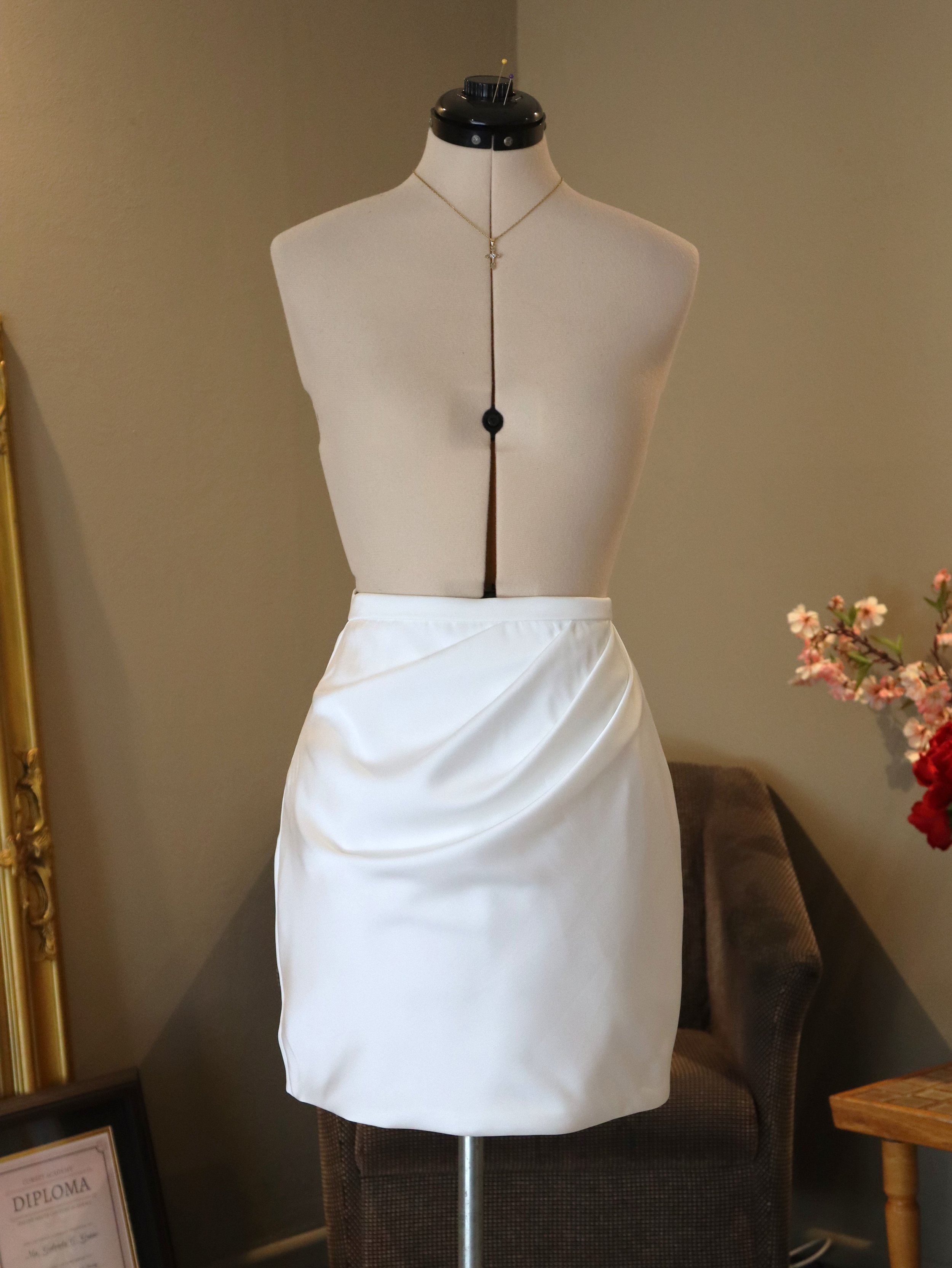 A mannequin displays a white satin skirt and a necklace with a locket on a beige background.