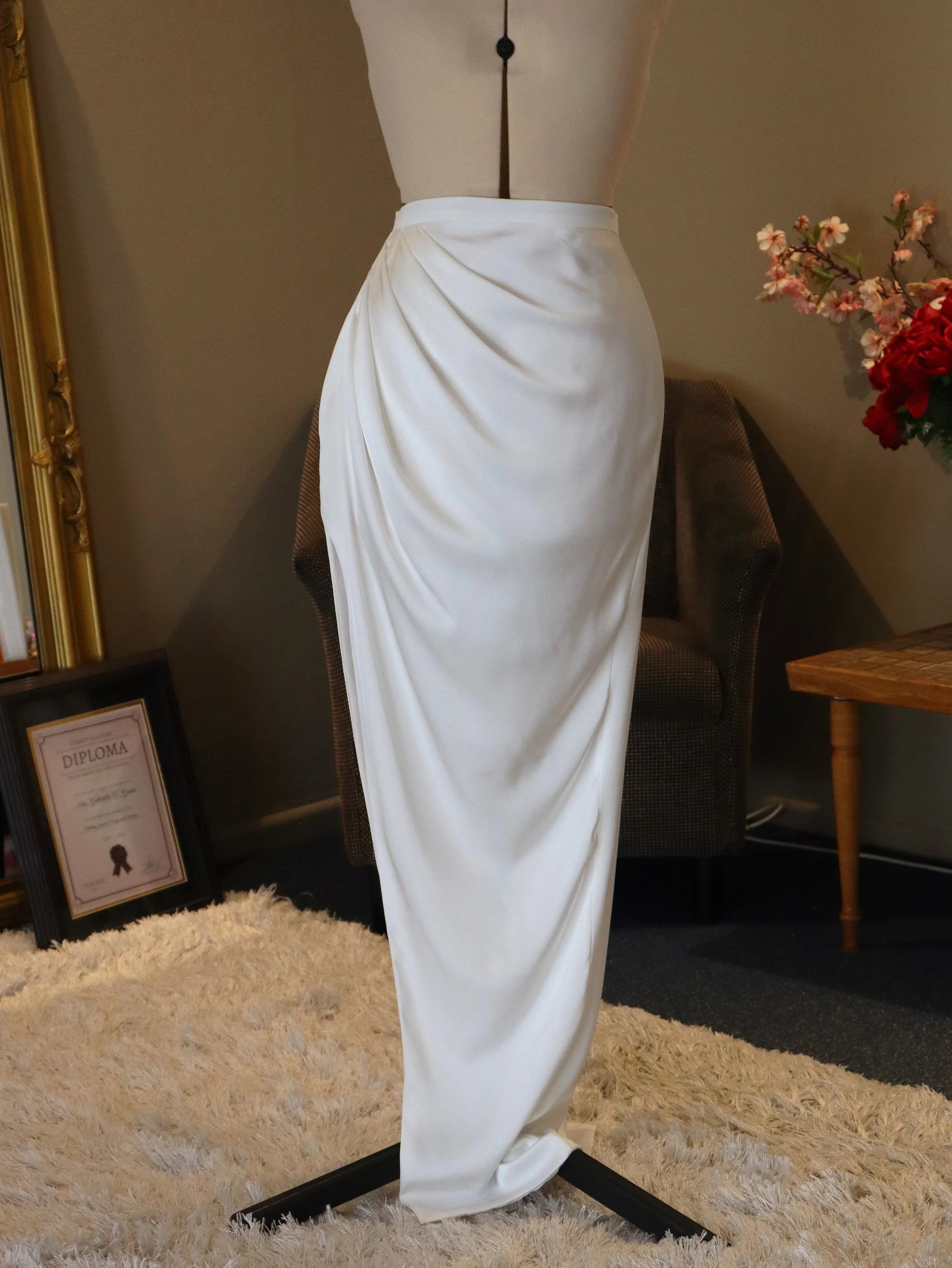 White satin skirt displayed on a dress form in a room with a beige carpet, velvet armchair, floral arrangement, and framed diploma.