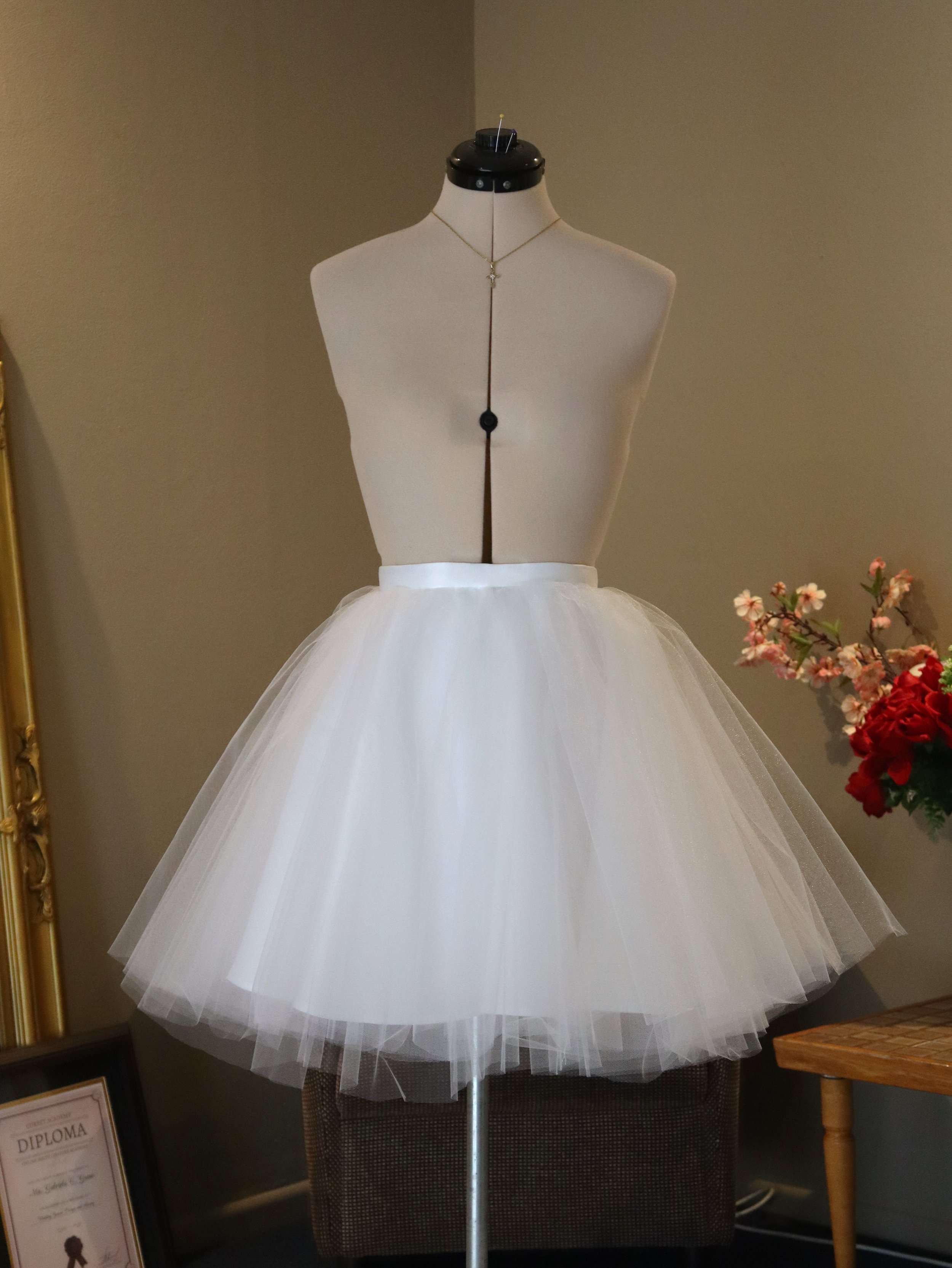 A mannequin dressed in a white tulle ballet tutu and a long, delicate necklace, displayed on a stand in a room with flowers and framed certificates.