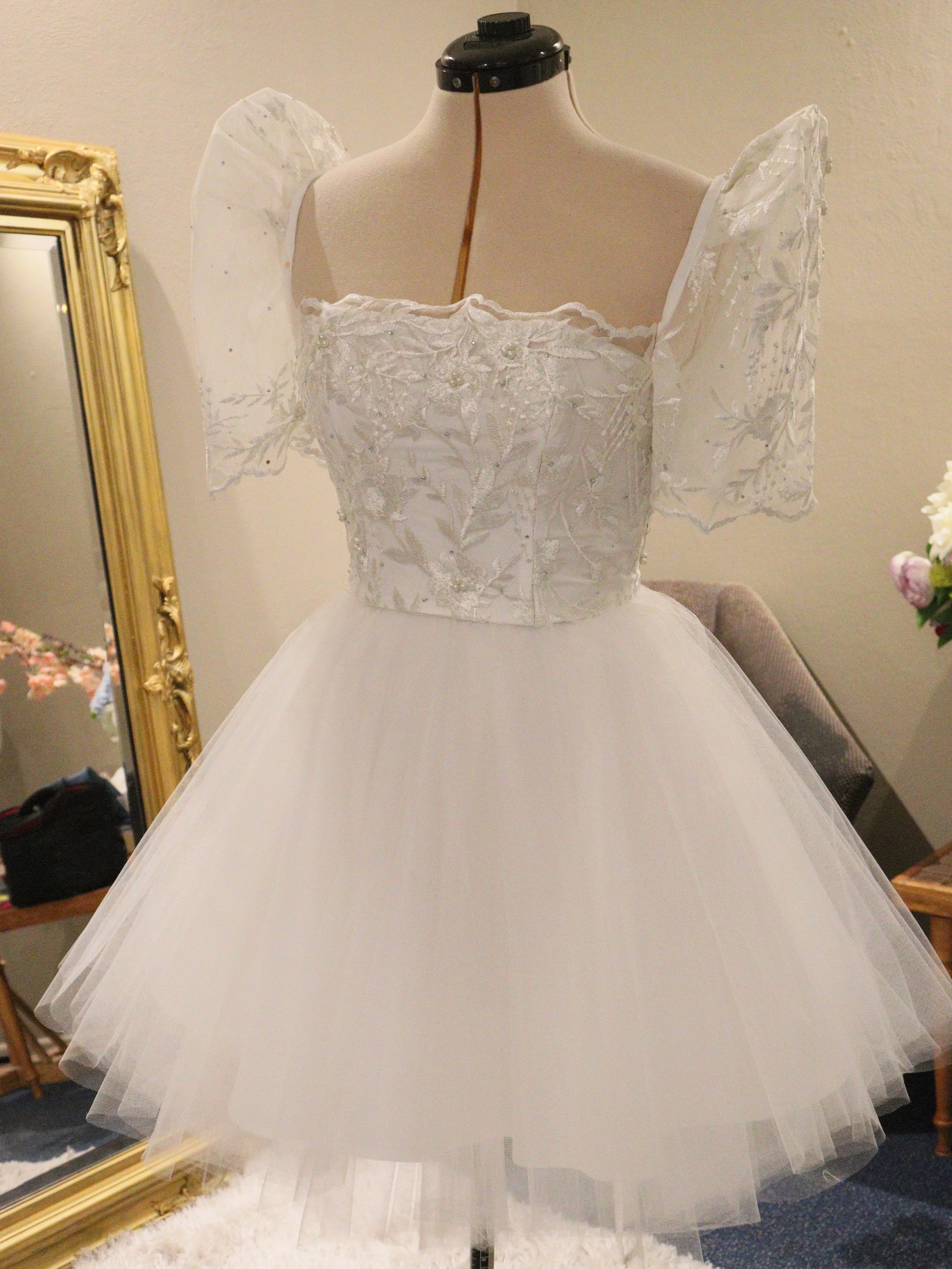 White wedding dress with embroidered bodice, puffed sleeves, and a full tulle skirt displayed on a dress form.