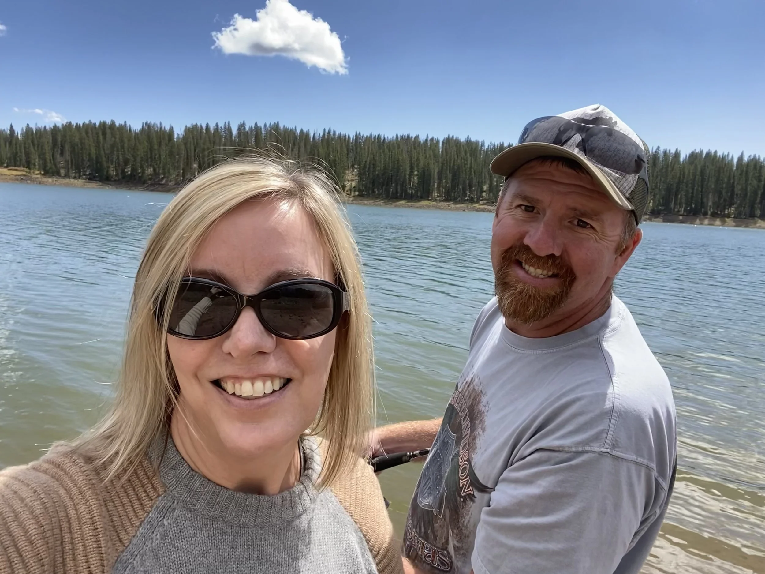 A smiling couple taking a selfie near a lake, with a woman wearing sunglasses and a beige sweater, and a man wearing a baseball cap and a gray t-shirt, standing with water and pine trees in the background.