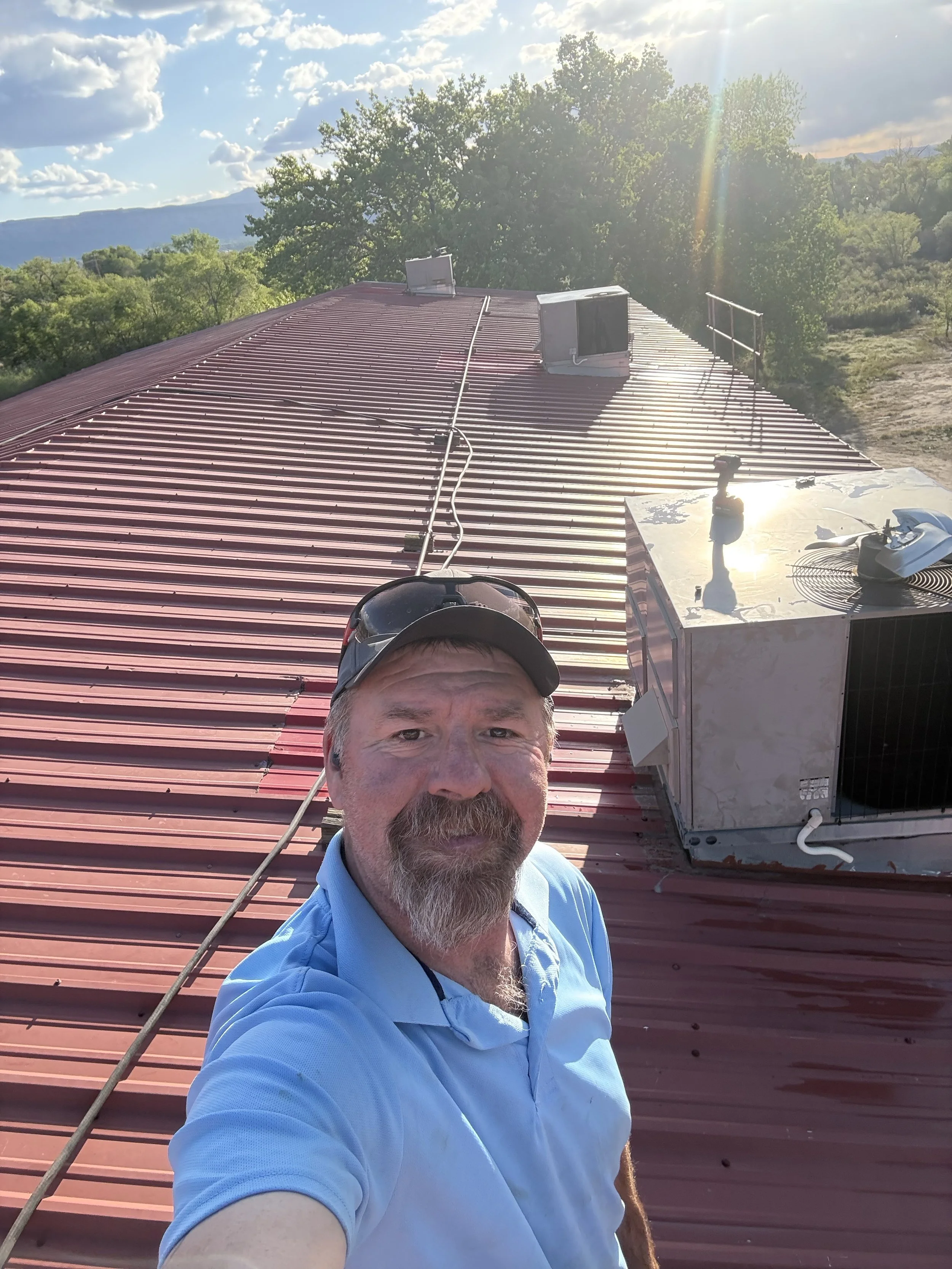 Self-portrait of a man wearing sunglasses and a blue collared shirt taking a selfie on a church rooftop with a red metal roof, HVAC units, green trees, mountains in the background, and a bright sunny sky in Grand Junction Colorado.