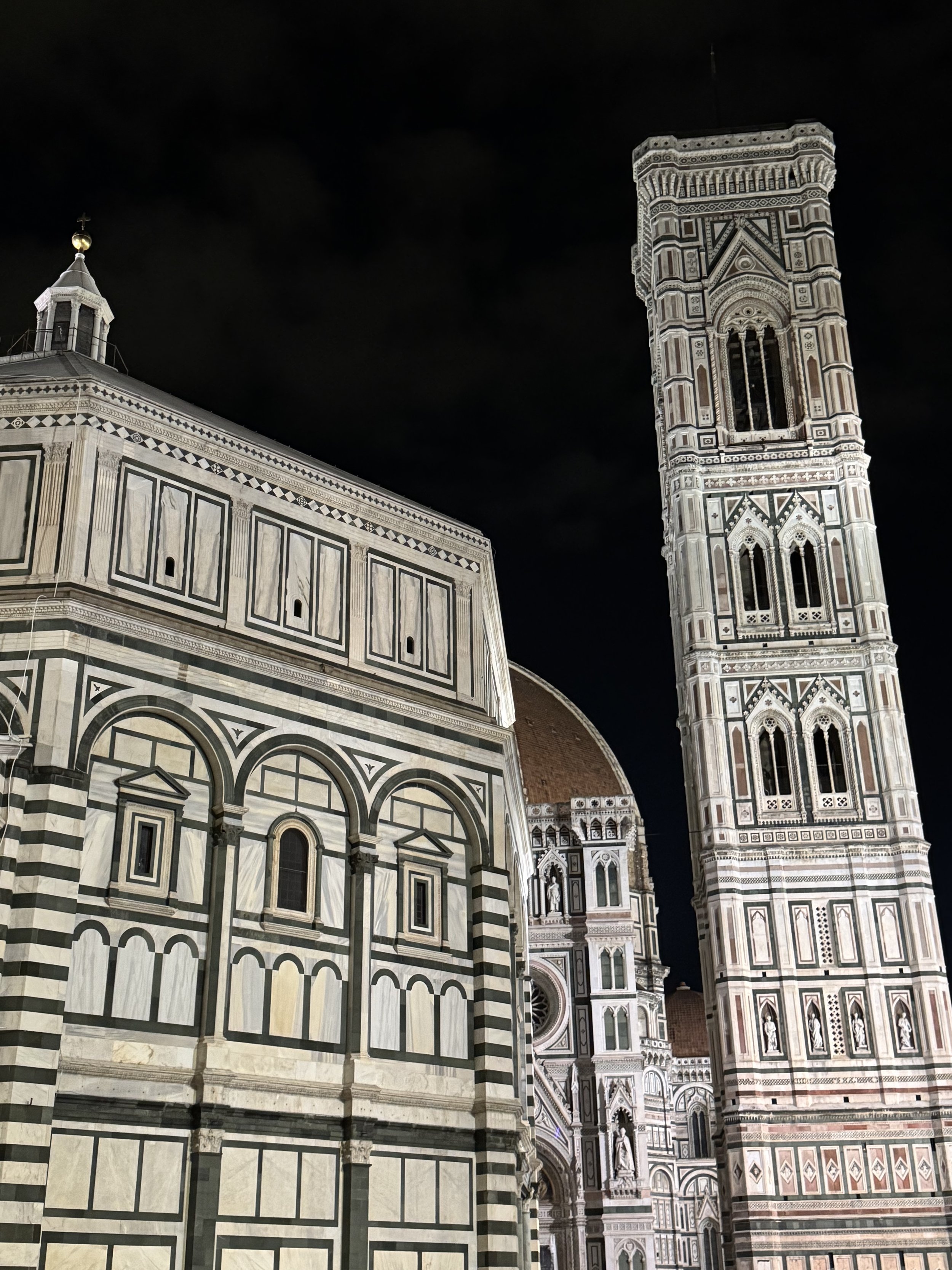 Night view of the Florence Cathedral, including the bell tower and the cathedral's ornate marble facade, illuminated against the dark sky.