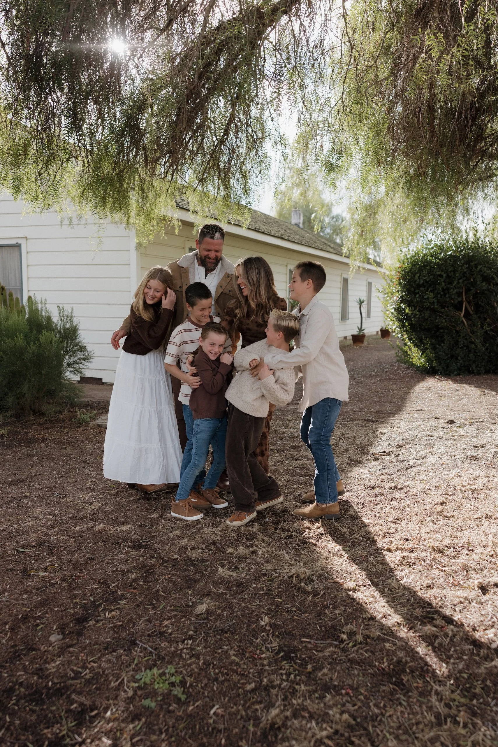 A family of seven, including adults and children, standing outdoors under a large tree with sunlight shining through the branches. They are smiling and enjoying a moment together near a white house with potted plants around.