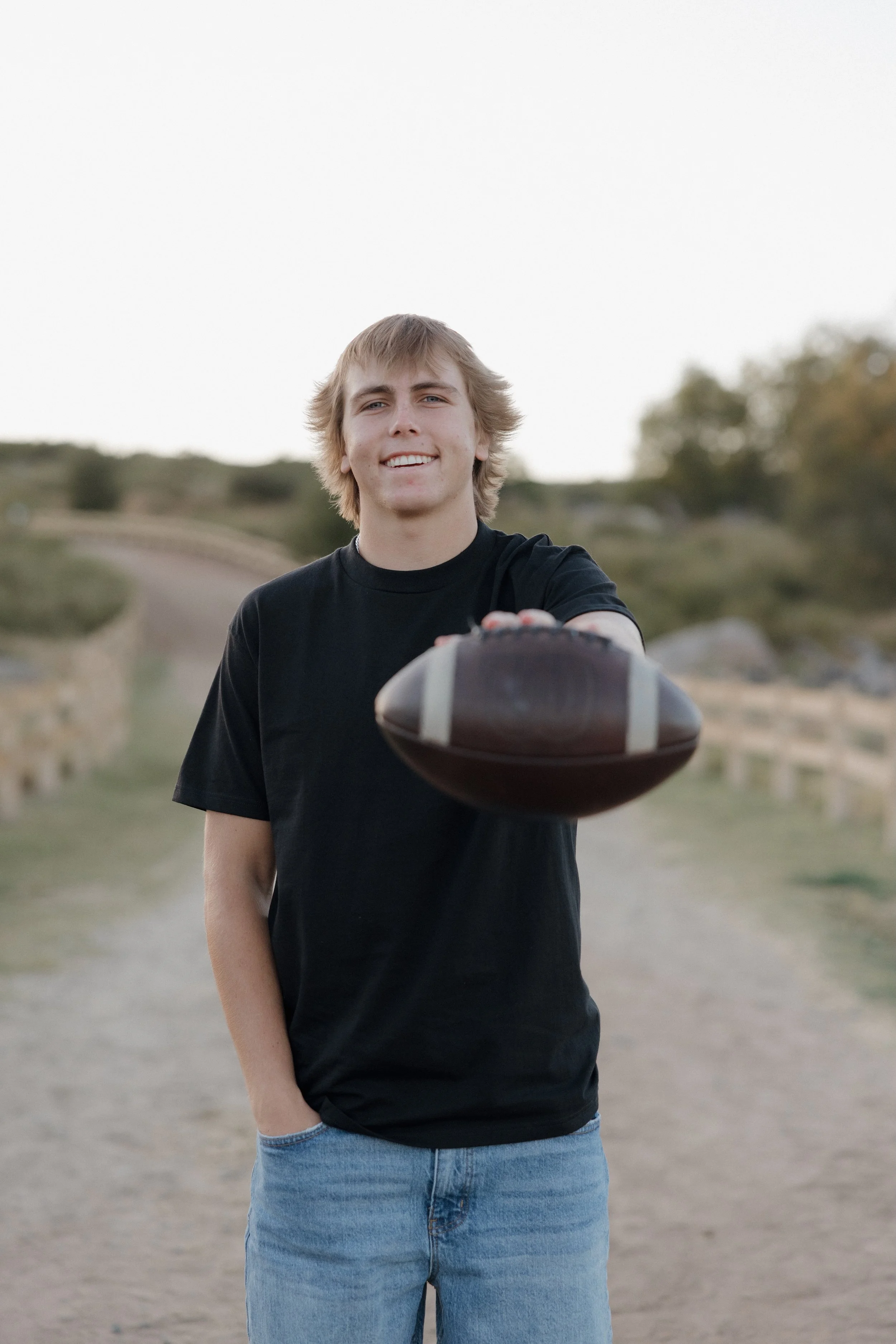 A young man with blond hair wearing a black T-shirt and blue jeans standing outdoors on a dirt path, extending his hand with a football.