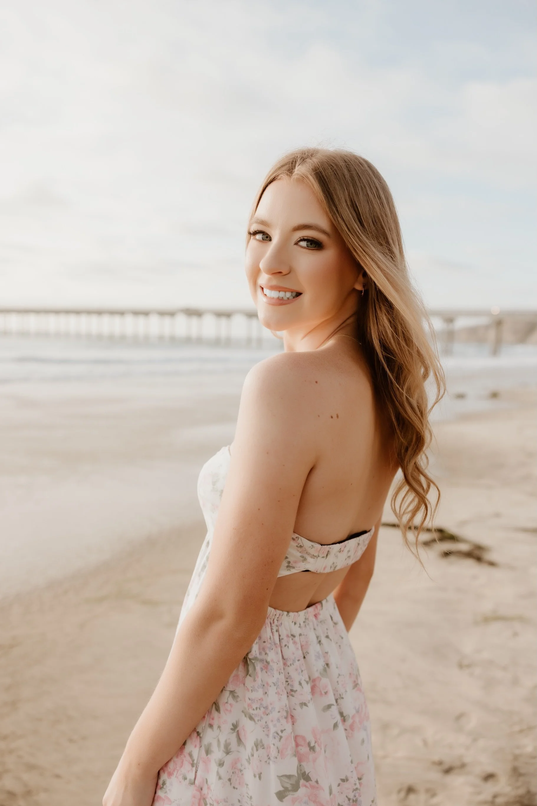 A young woman with long wavy hair smiling at the camera on a beach with a pier in the background.