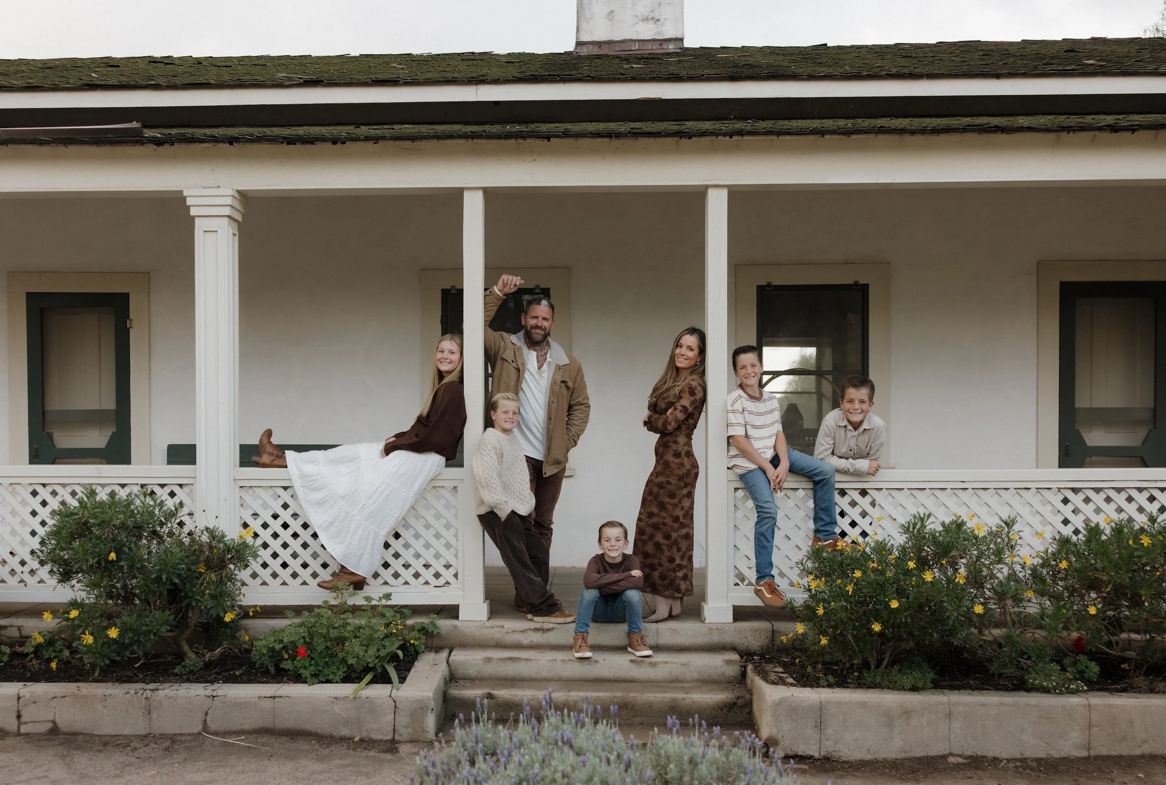 Family of seven posing on front porch of a house, with three children sitting on the steps, two standing, and two adults standing behind them. The house has a white exterior, green door, and a porch railing with lattice. There are plants and flowers in the garden in front.