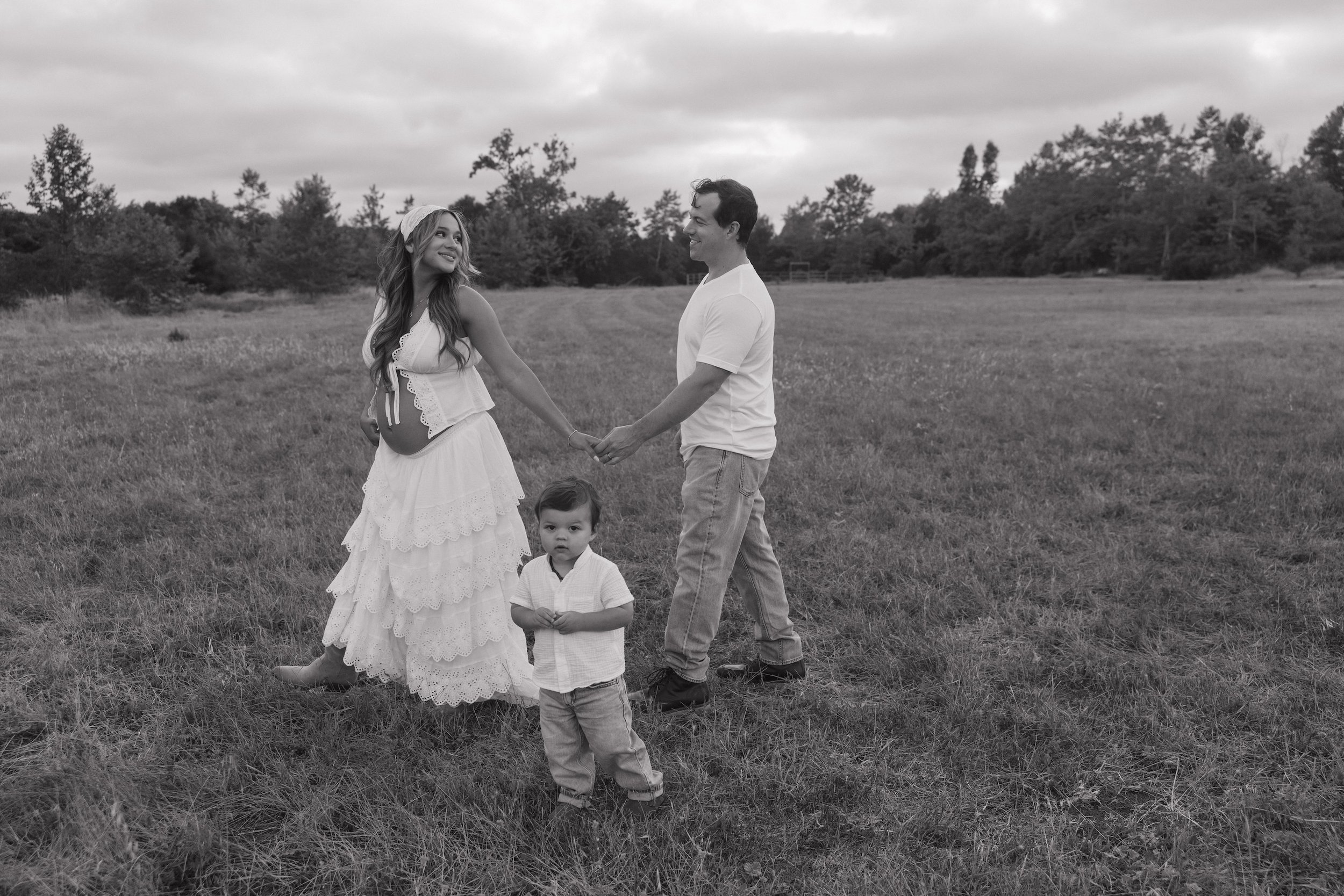 A black and white photo of a family of four standing in a grassy field, holding hands. The woman is pregnant and smiling at a man, while two young children stand nearby, one looking at the camera and the other holding hands with the woman.